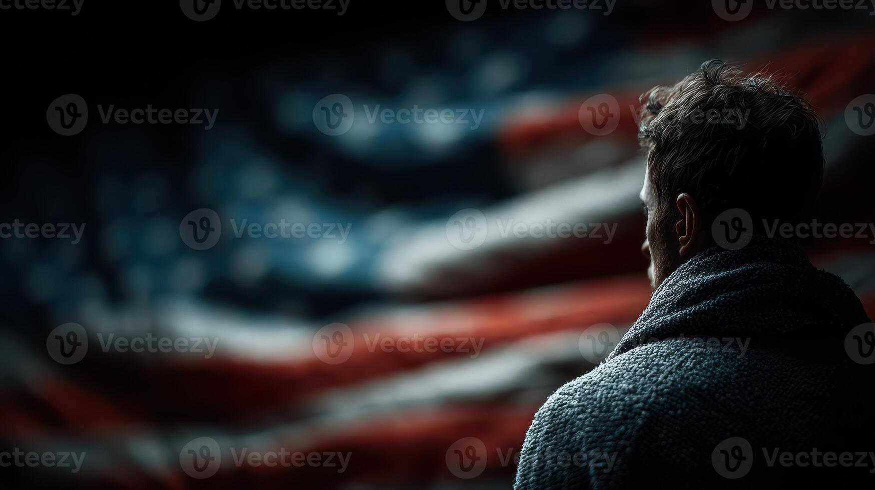 Man gazes thoughtfully towards a large American flag in a darkened environment, showcasing a moment of reflection and patriotism photo