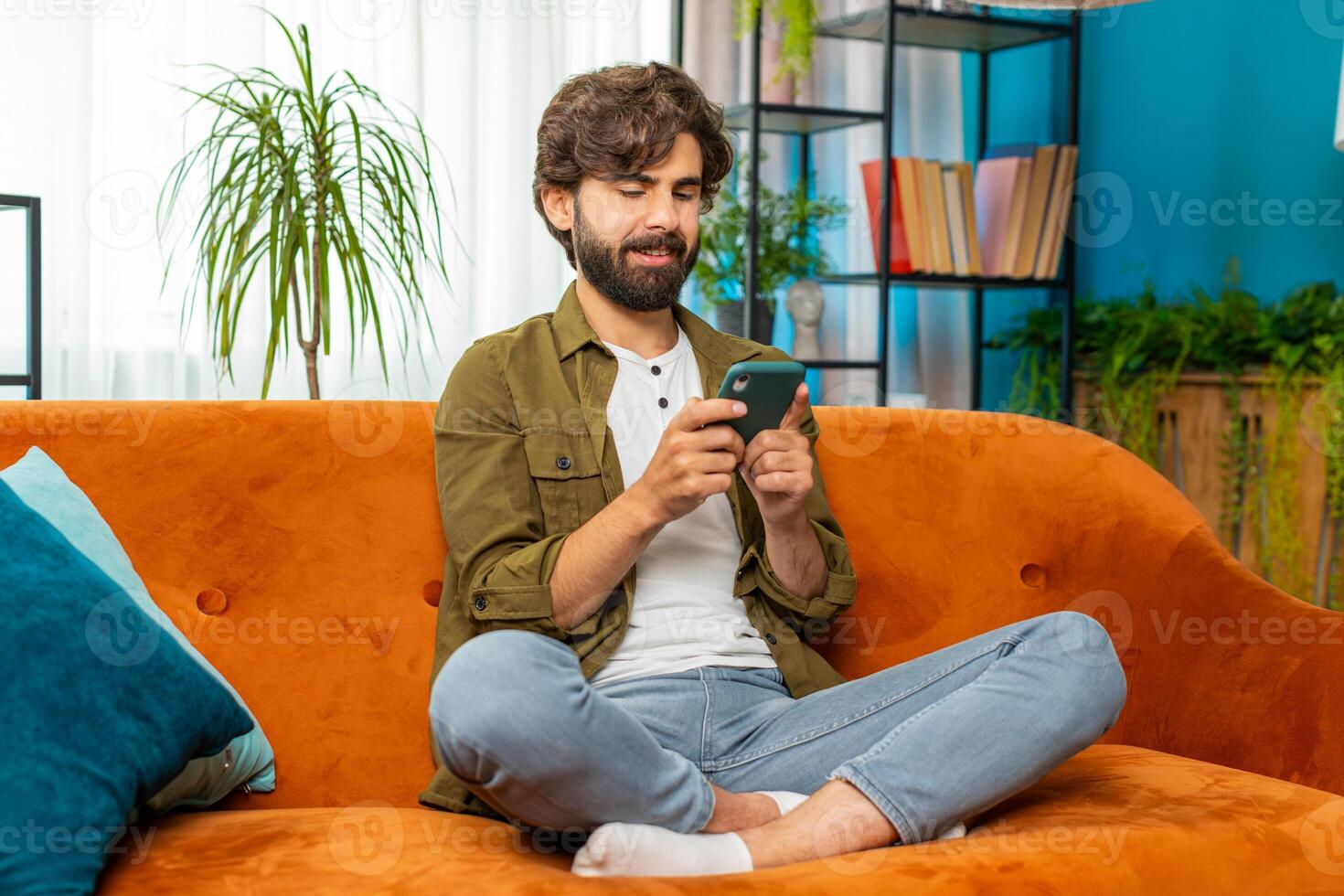 Middle eastern young man sitting on sofa using smartphone share messages on social media application photo