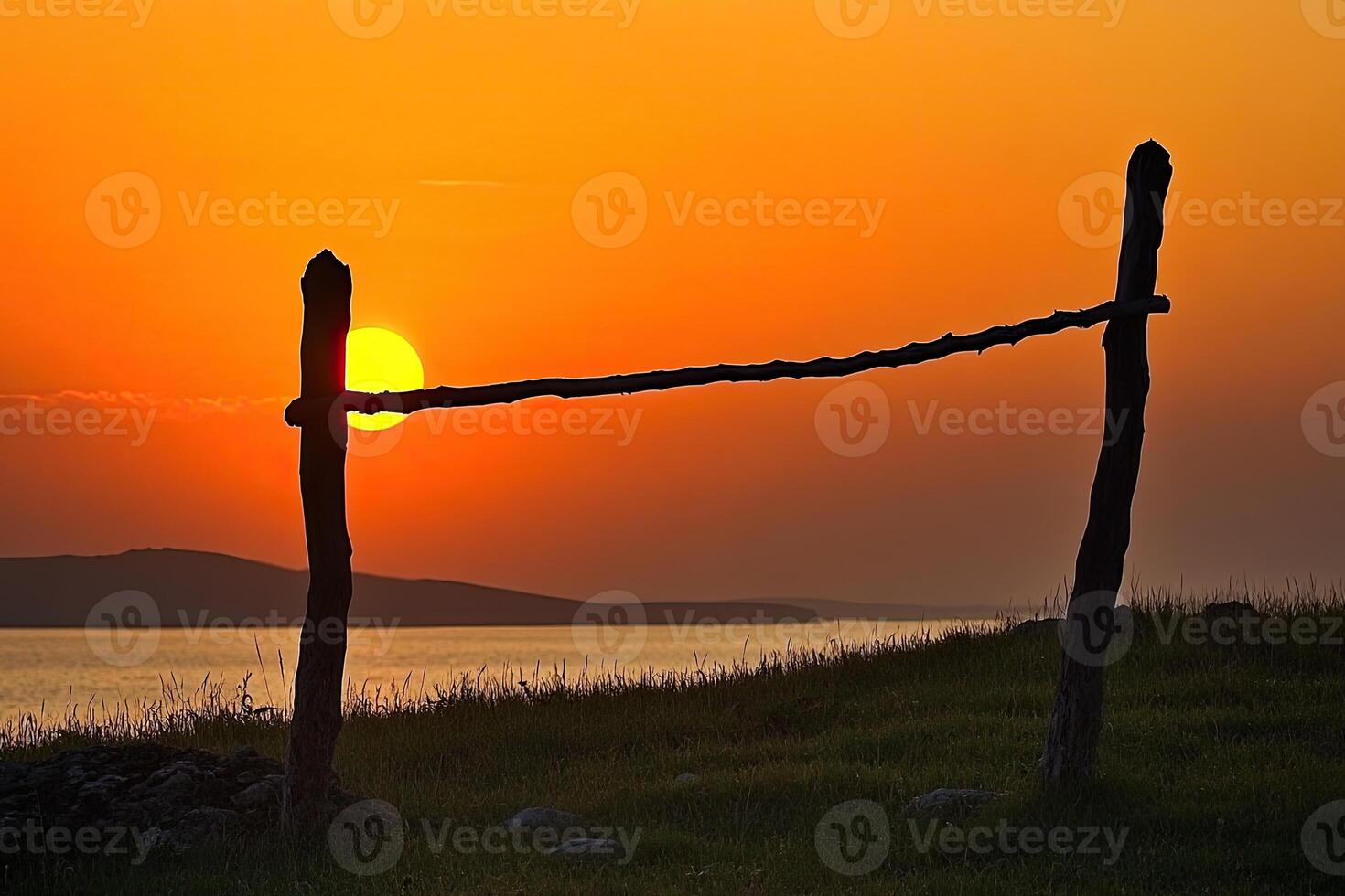 Sunset silhouettes of simple wooden posts and a rustic fence against a vibrant orange sky over a calm body of water photo