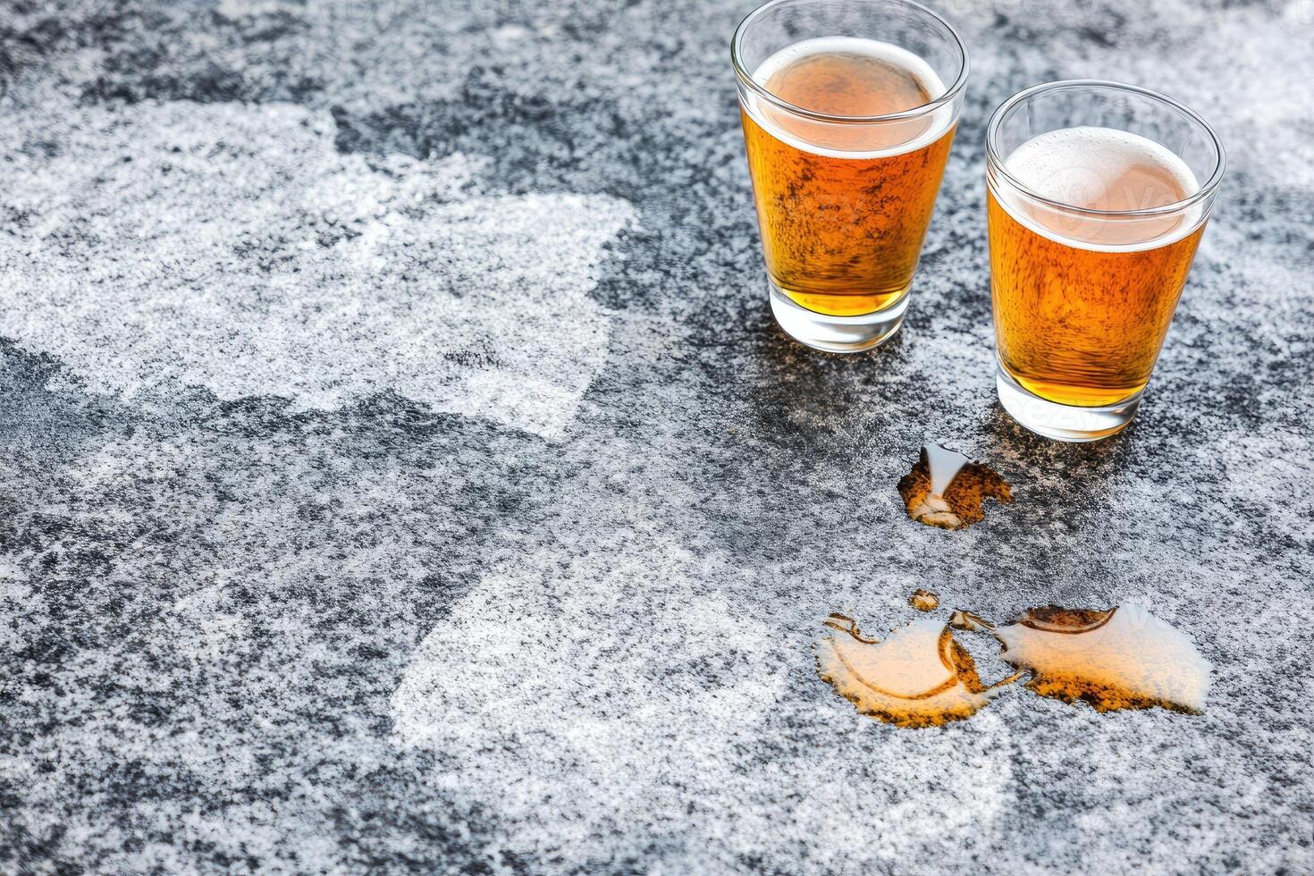 Two glasses of light amber beer on a mottled gray surface with spilled beer photo