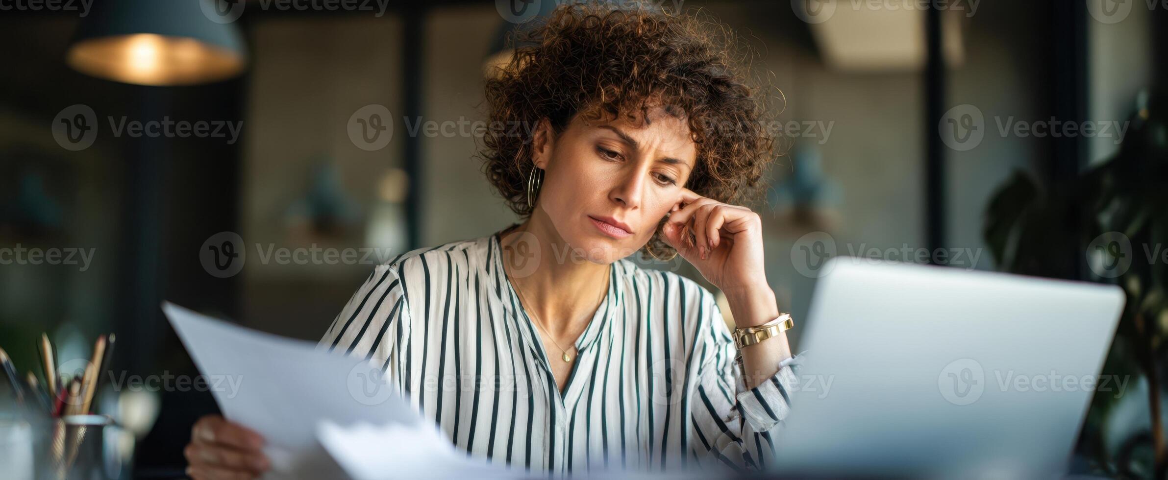 The woman thoughtfully analyzes documents while seated at her modern office workspace. photo