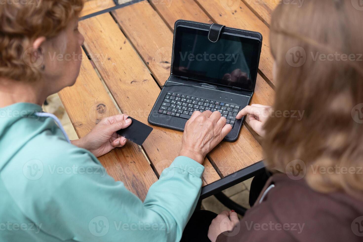 A grandma and her granddaughter are at an outdoor table with a laptop, the grandma is holding a credit card and celebrating, suggesting they successfully made a transaction. photo