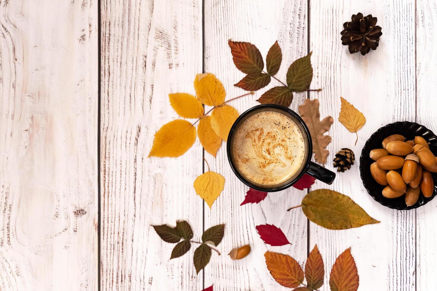Overhead shot of an autumn-themed arrangement with a white coffee cup and saucer as the central element, complemented by fallen leaves, acorns, and a delicate floral accent. photo