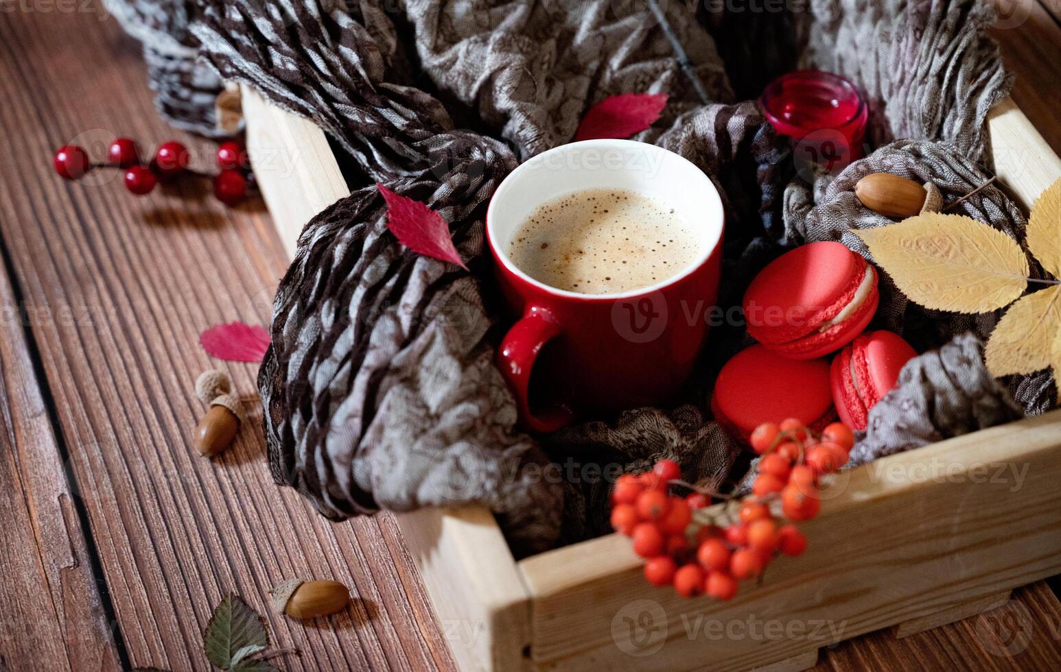 A close-up, overhead shot of a wooden box filled with a textured grey scarf, a red-handled mug of coffee, red macarons, rowan berries, a yellow leaf, and an acorn. photo