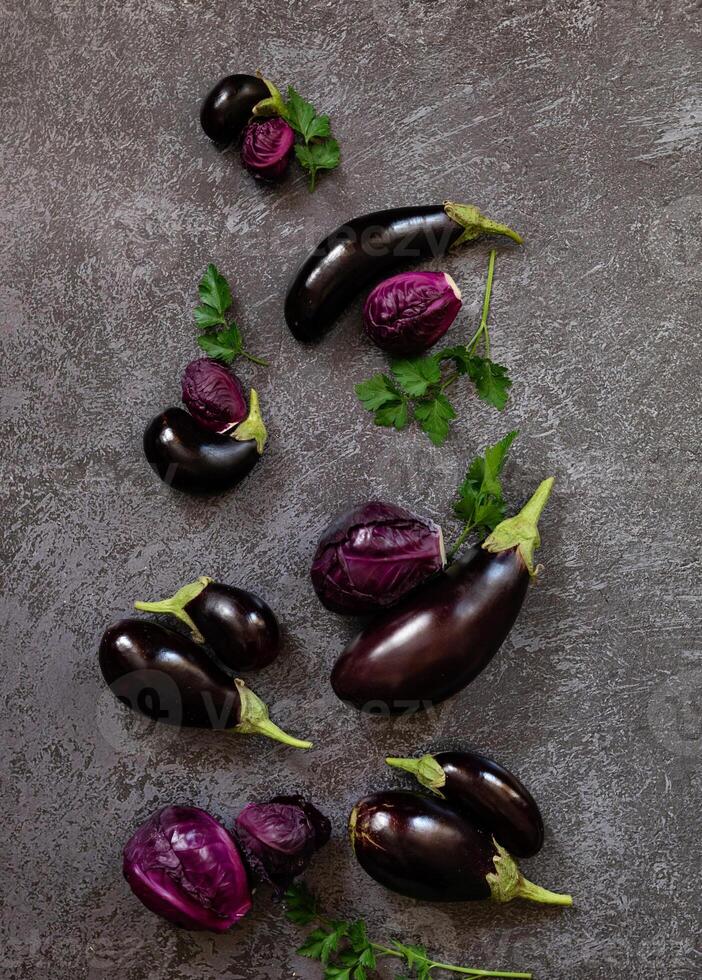 Close-up of a table arrangement with various eggplants and fresh parsley, with a knife placed on a textured, grey background. photo