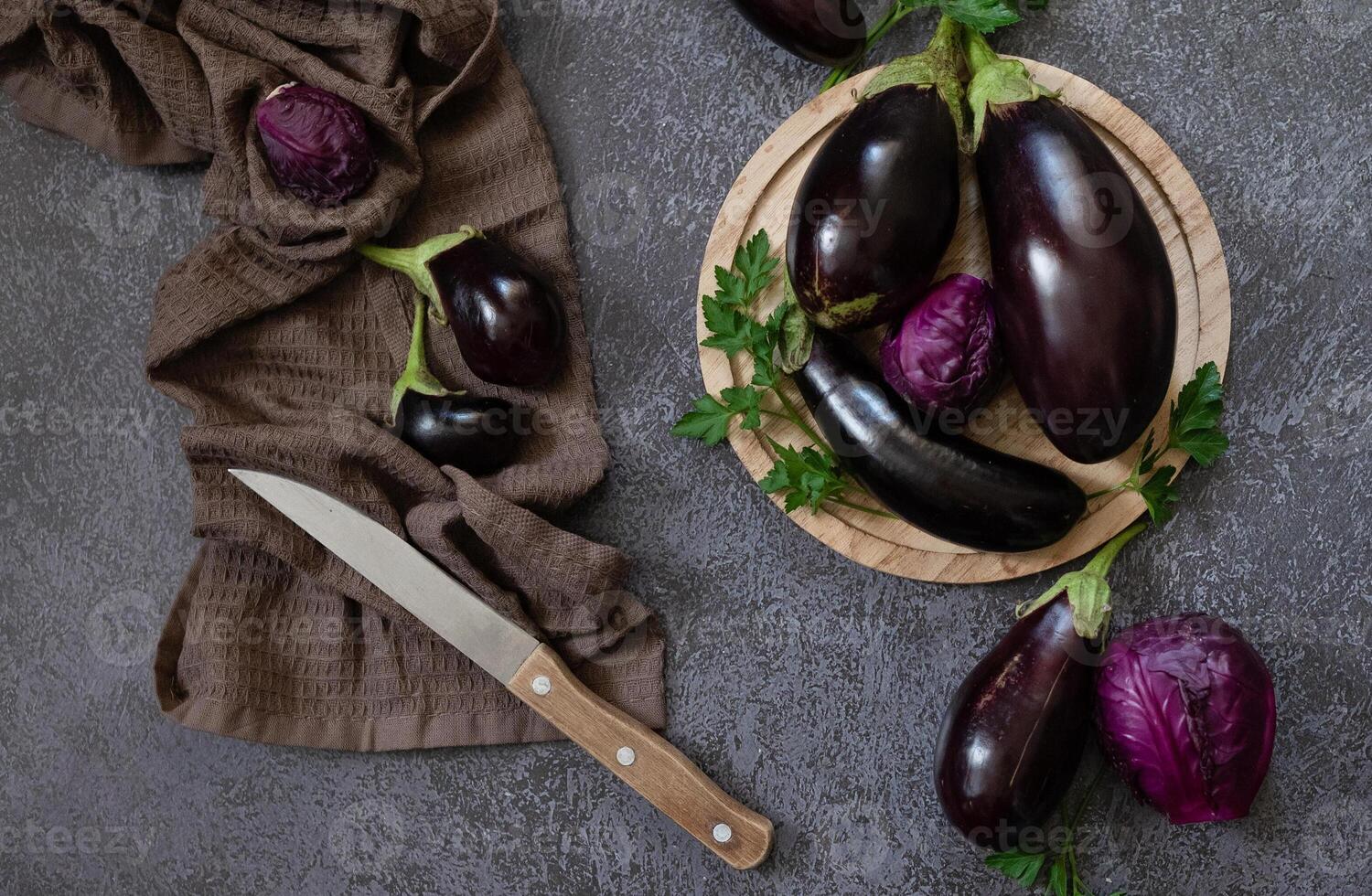 A photograph of a culinary setting with eggplants and parsley, with a knife, set against a textured, grey background. photo