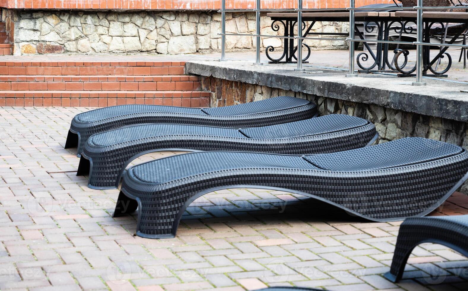 The image shows a row of modern black wicker lounge chairs neatly arranged on a paved patio area next to a stone and brick wall. photo