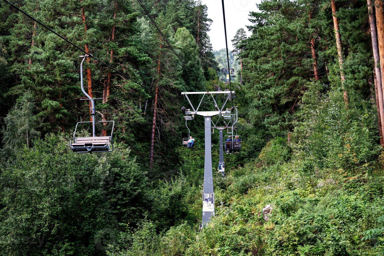 Chairlifts carry passengers over a dense, green forest with tall trees under a cloudy sky. photo