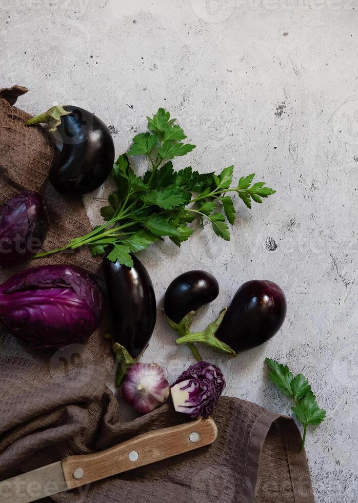 A flat lay image featuring eggplants, parsley, and a knife on a textured, grey surface. photo