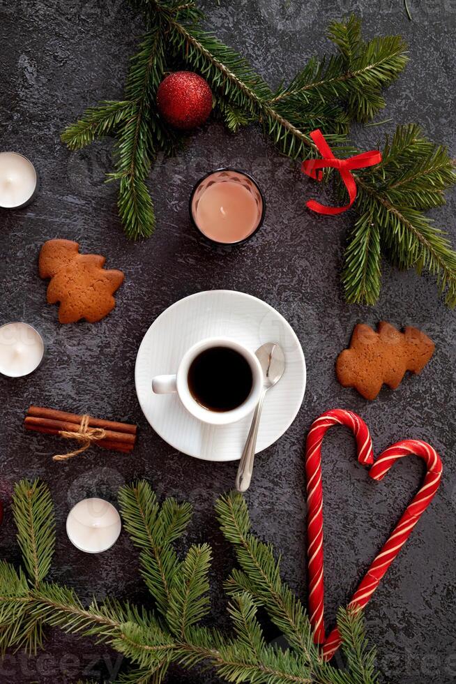 A seasonal still life highlighting a coffee cup, accompanied by gingerbread cookies, cinnamon sticks, candy canes, candles, and pine sprigs, all placed strategically on a dark table. photo