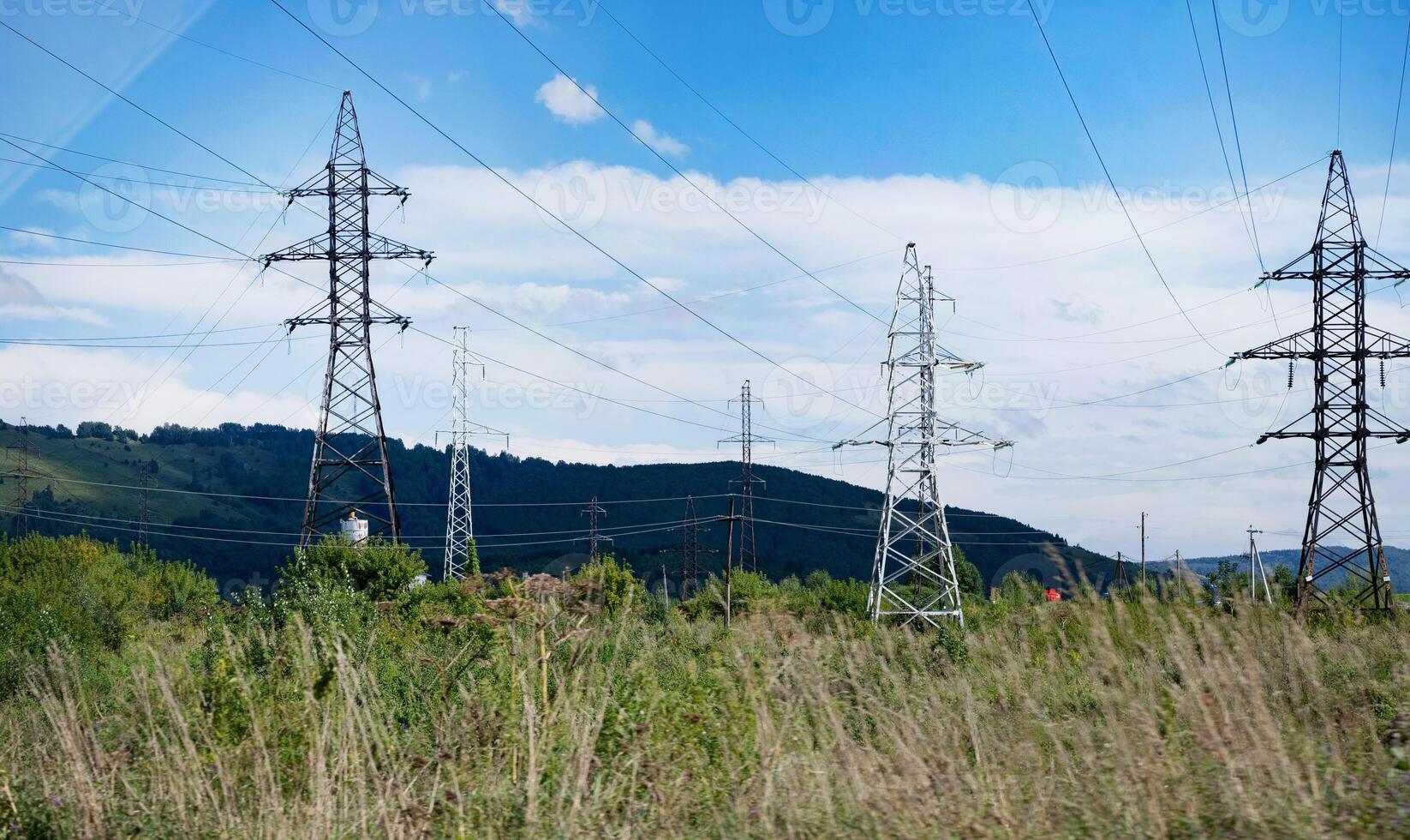 The image shows several tall electrical transmission towers standing in a grassy field with hills in the background under a partly cloudy blue sky. photo