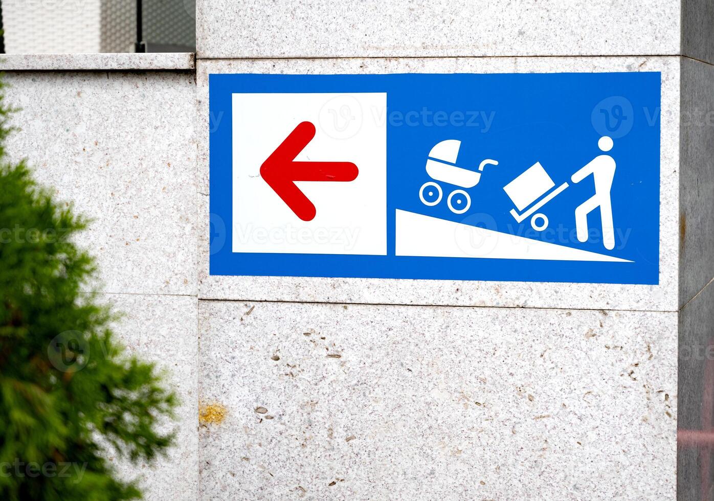 Sign with a red left arrow and icons showing a person with a cart and stroller on a ramp. Placed on a stone wall background. Accessibility concept photo