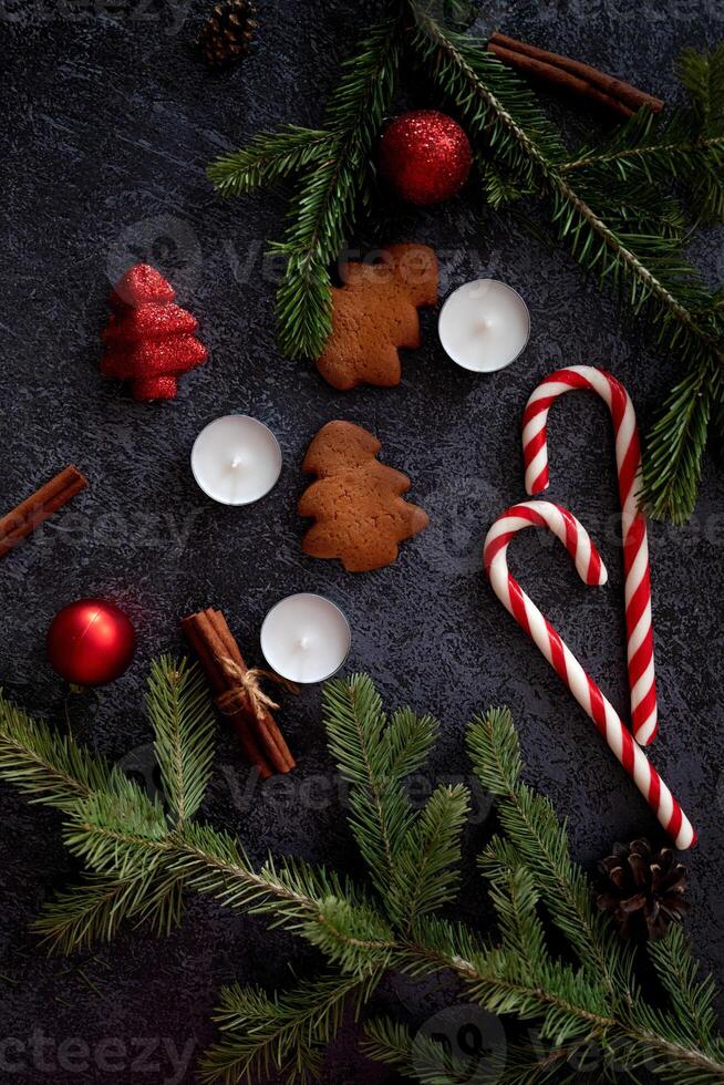 An aesthetic Christmas flat lay composition on a dark backdrop, highlighting a cup of coffee, gingerbread cookies, cinnamon sticks, candy canes, candles, and festive pine sprigs. photo