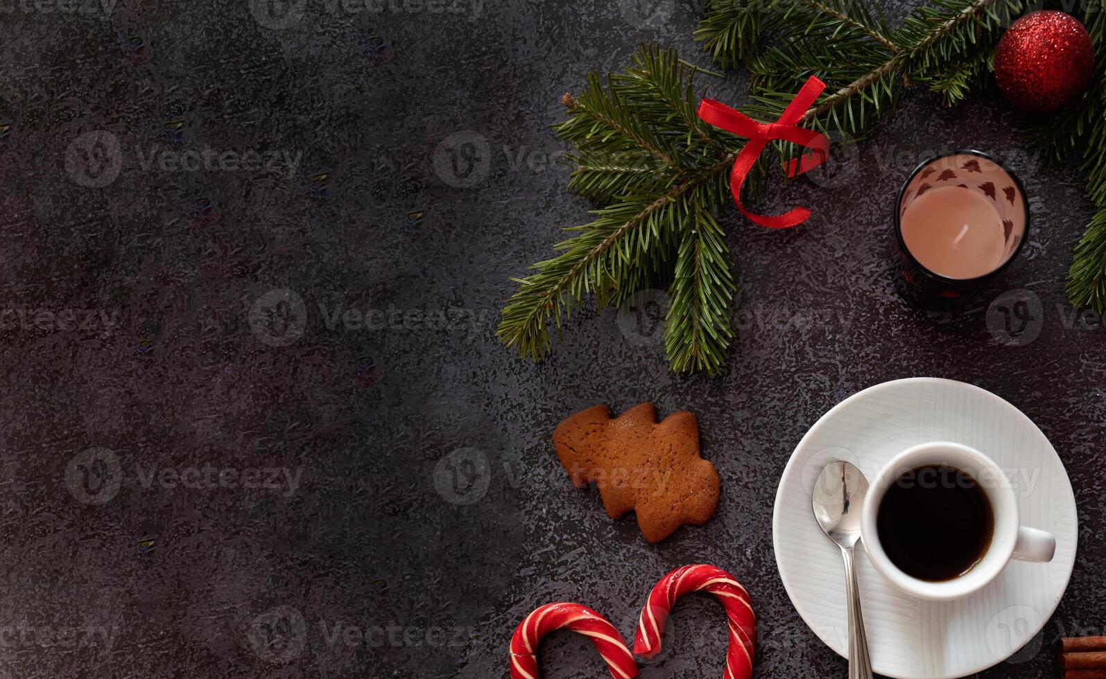 An artistic top shot of a Christmas decoration arrangement featuring coffee, gingerbread cookies, cinnamon sticks, candy canes, candles, and pine sprigs placed on a dark textured surface. photo
