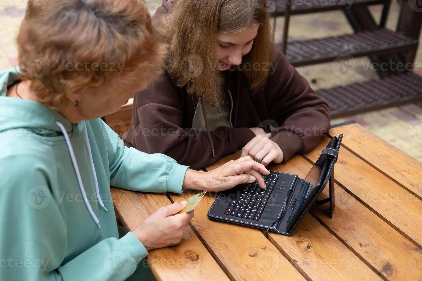 Two people are sitting at a wooden table, interacting with a small tablet device that has a keyboard attached. photo