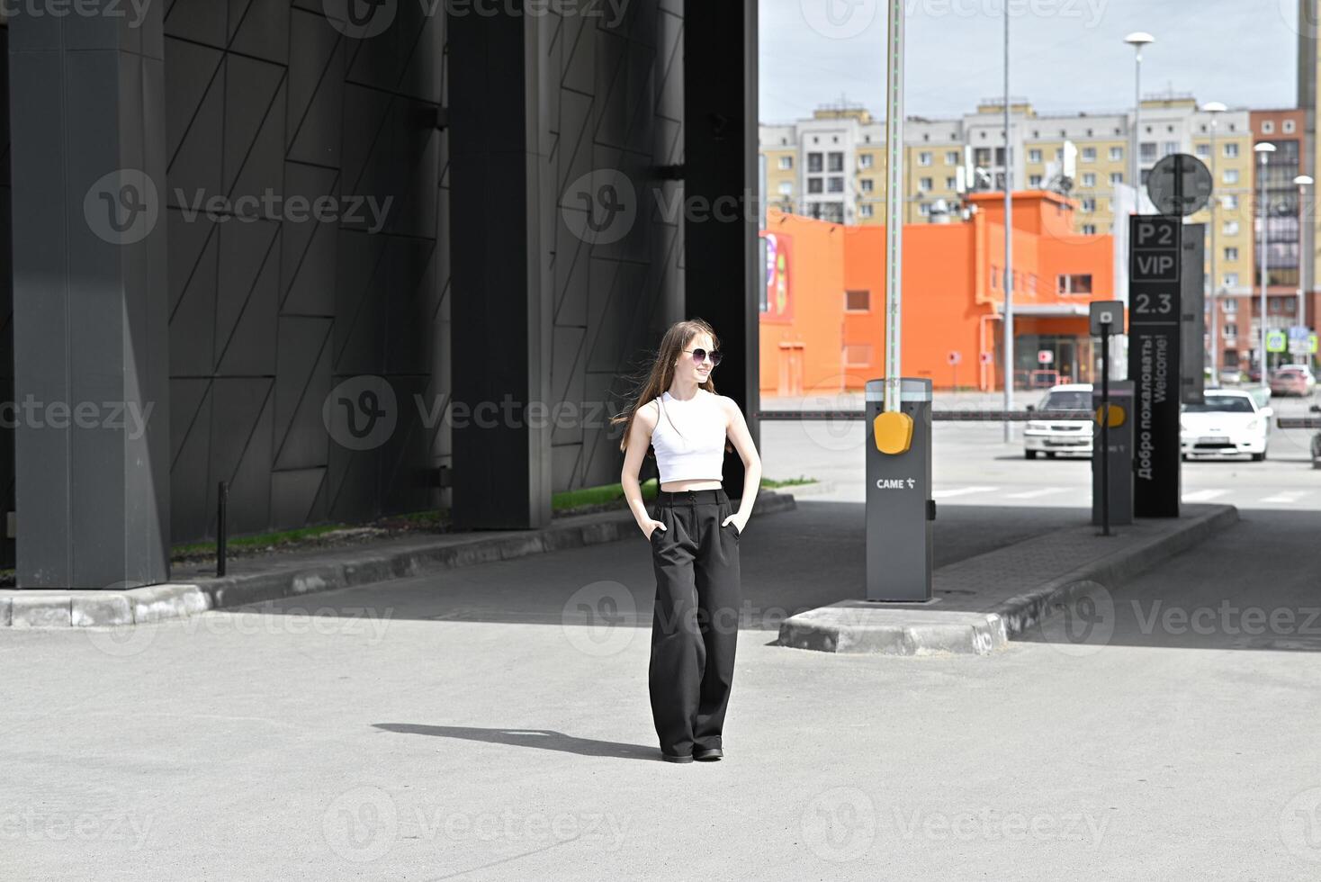 A solitary figure in fashionable black wide-leg pants and a white sleeveless top, standing on an empty paved area. photo