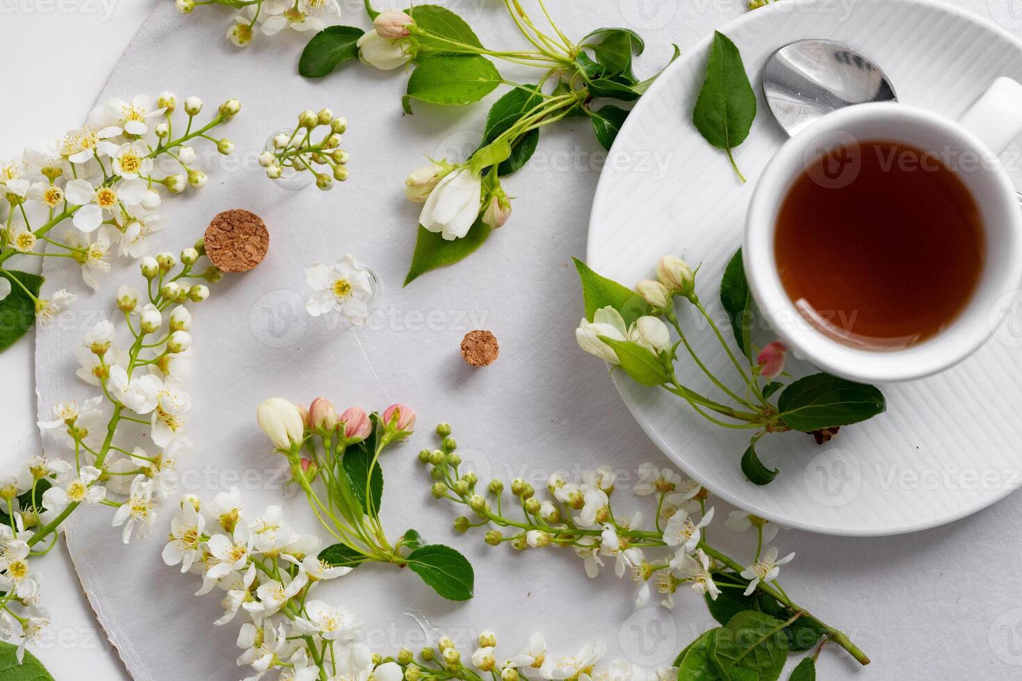 The image depicts a delicate arrangement of small white blossoms and green leaves scattered elegantly on a white surface, surrounding a white cup filled with tea placed on a matching saucer. photo