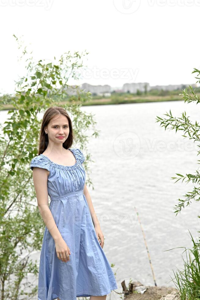 Young woman smiling by a lake in a blue dress with trees and water in the background. photo