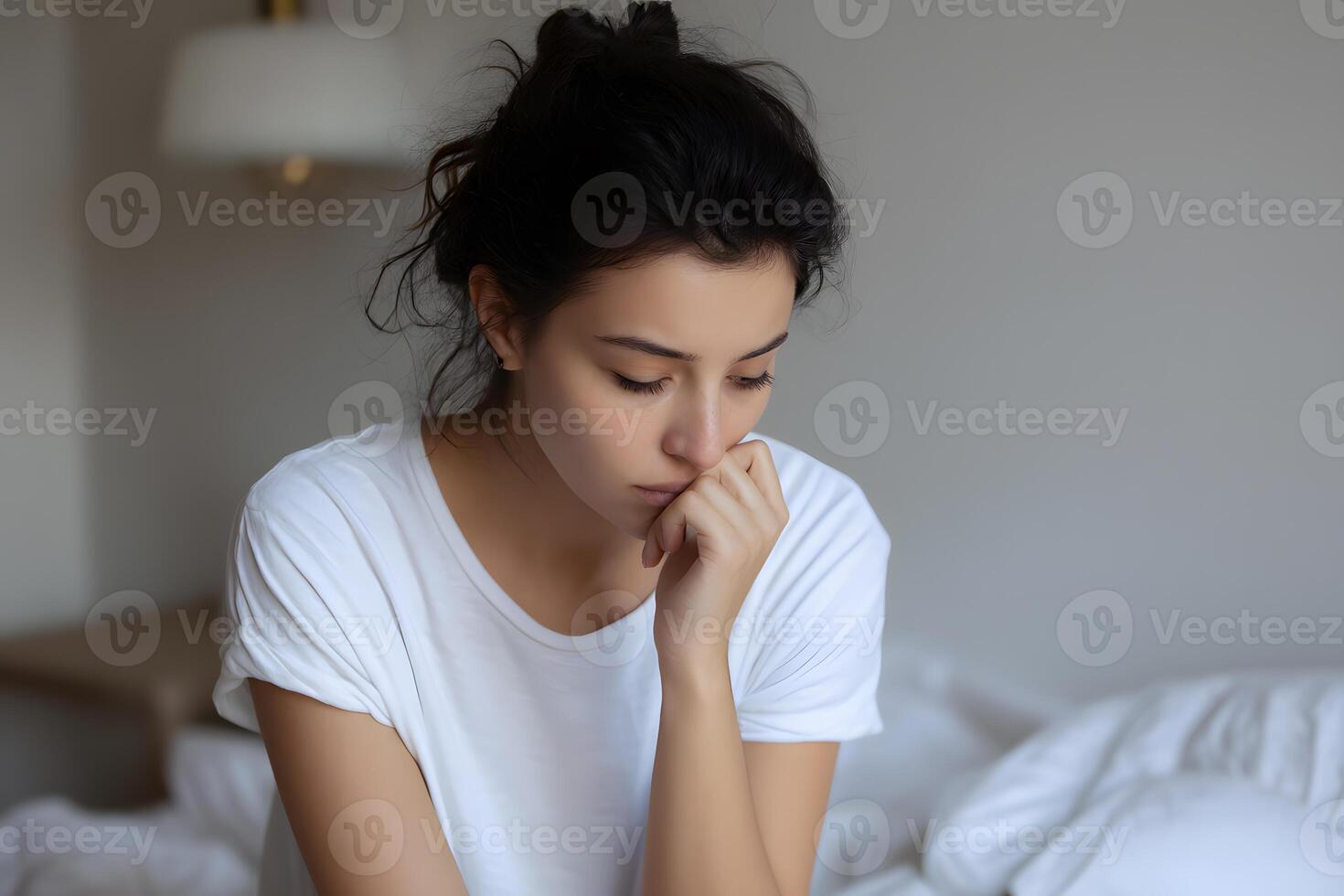Tired and anxious young woman in white bedding, head resting on hand, looking down in deep concern photo