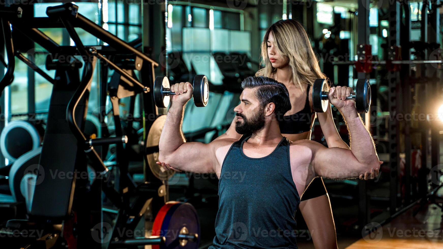 Personal trainer assisting man lifting dumbbells in gym, focusing on strength training, muscle building, and guided workout for fitness and health improvement. photo