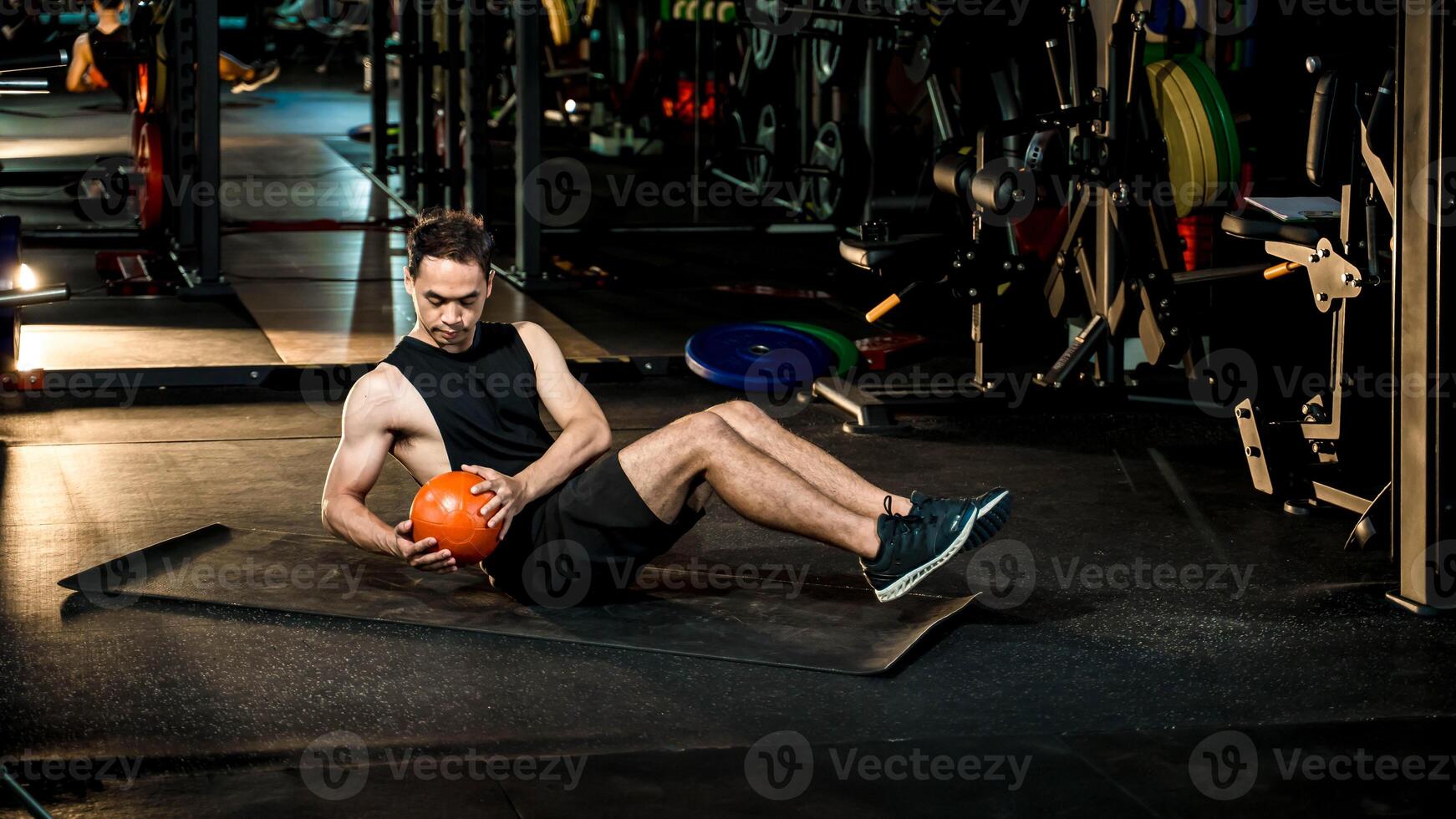 Focused young man performing Russian twist with medicine ball on yoga mat at gym, strengthening core and improving balance during functional workout session. photo