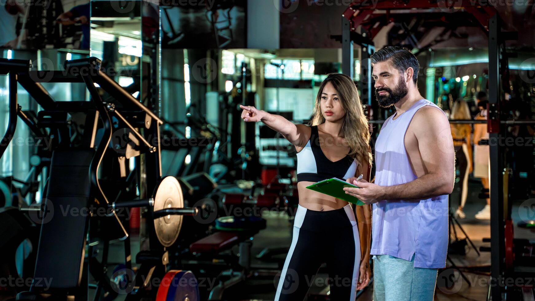 Female fitness coach pointing out training plan direction to male client at gym, holding clipboard while explaining workout steps during personal training session photo