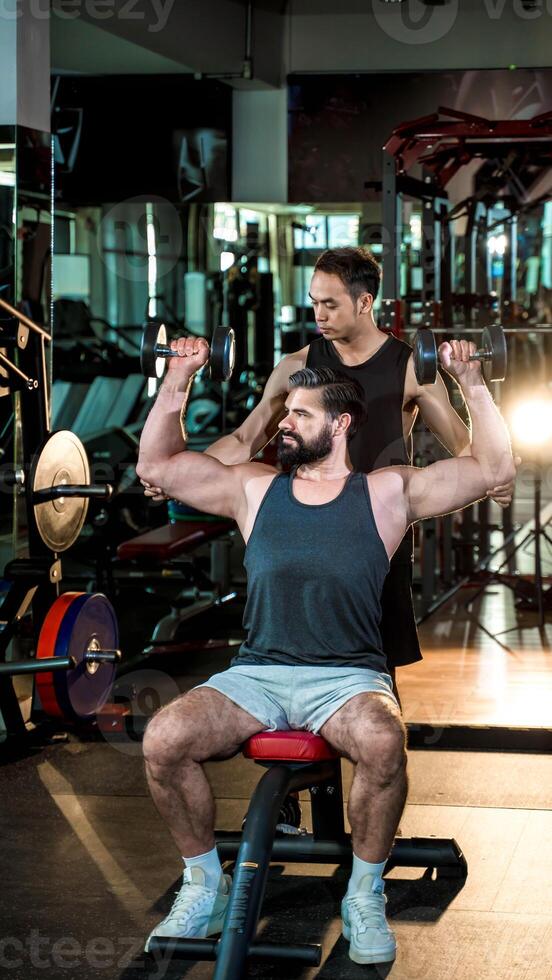 Personal trainer guiding man in seated dumbbell shoulder press exercise at gym, focusing on muscle building, strength training, and fitness motivation. photo