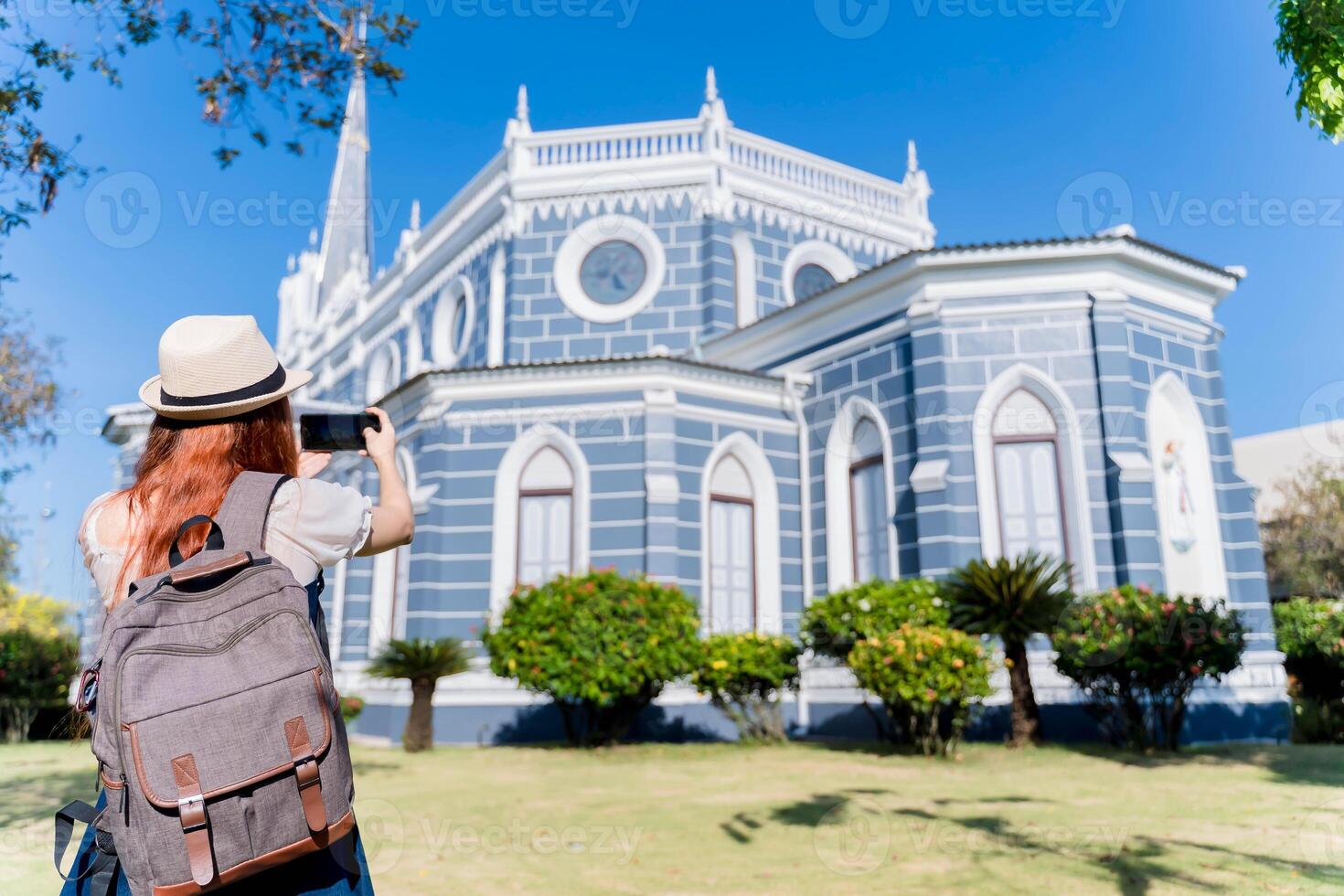 Young female tourist with backpack and hat photographing a blue historic cathedral on a sunny day, capturing architectural beauty during solo travel adventure photo