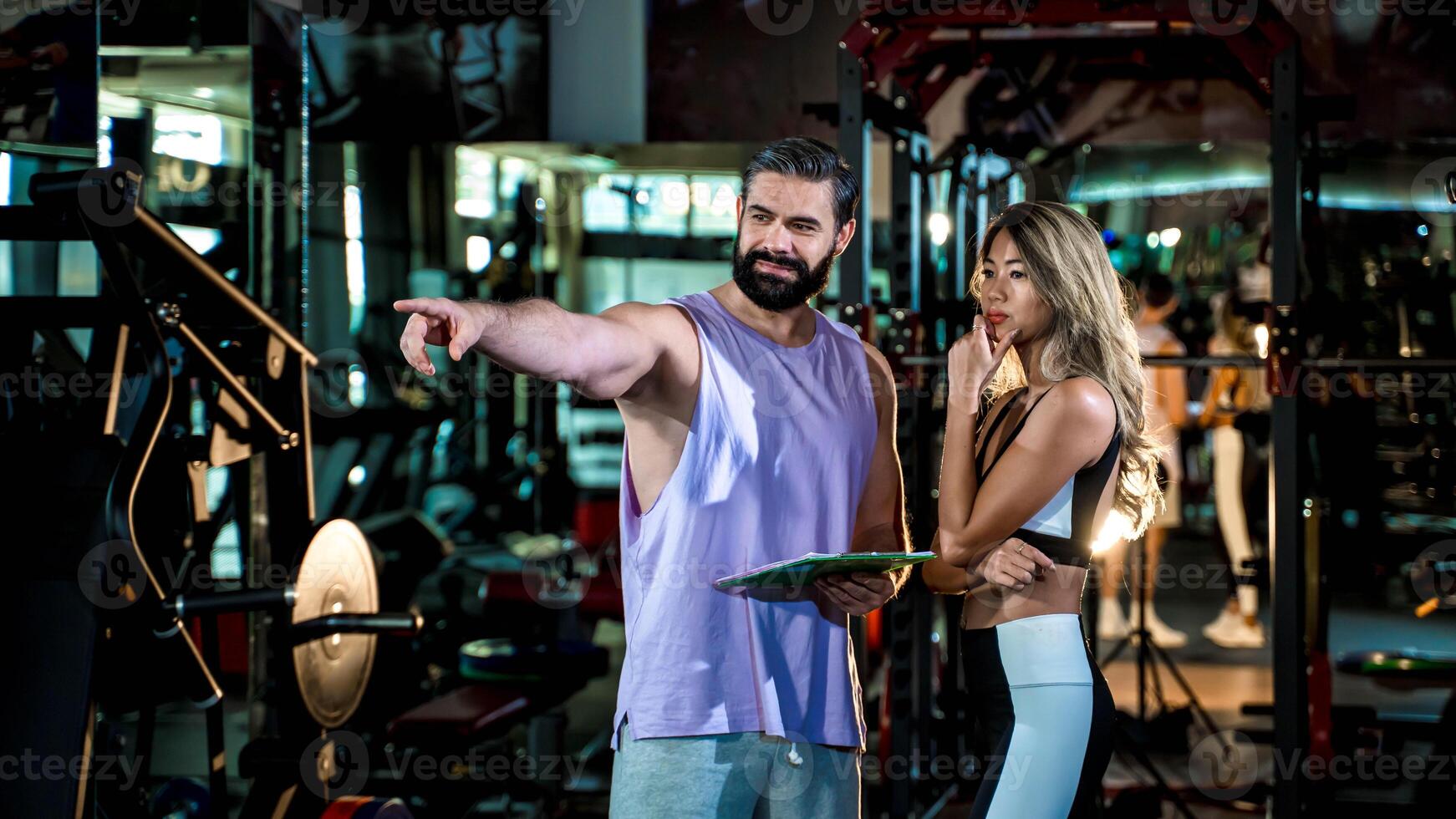 Personal trainer discussing workout strategy with female client at the gym, pointing towards equipment while planning fitness goals with clipboard in hand photo