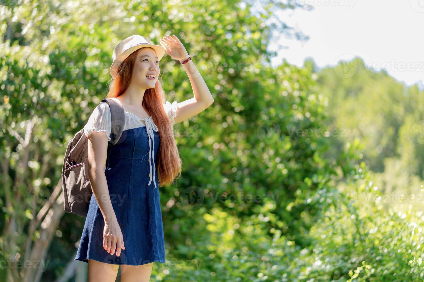 Happy young woman shielding eyes from the sun while hiking in a lush green forest, enjoying outdoor adventure and nature on a bright summer day. photo