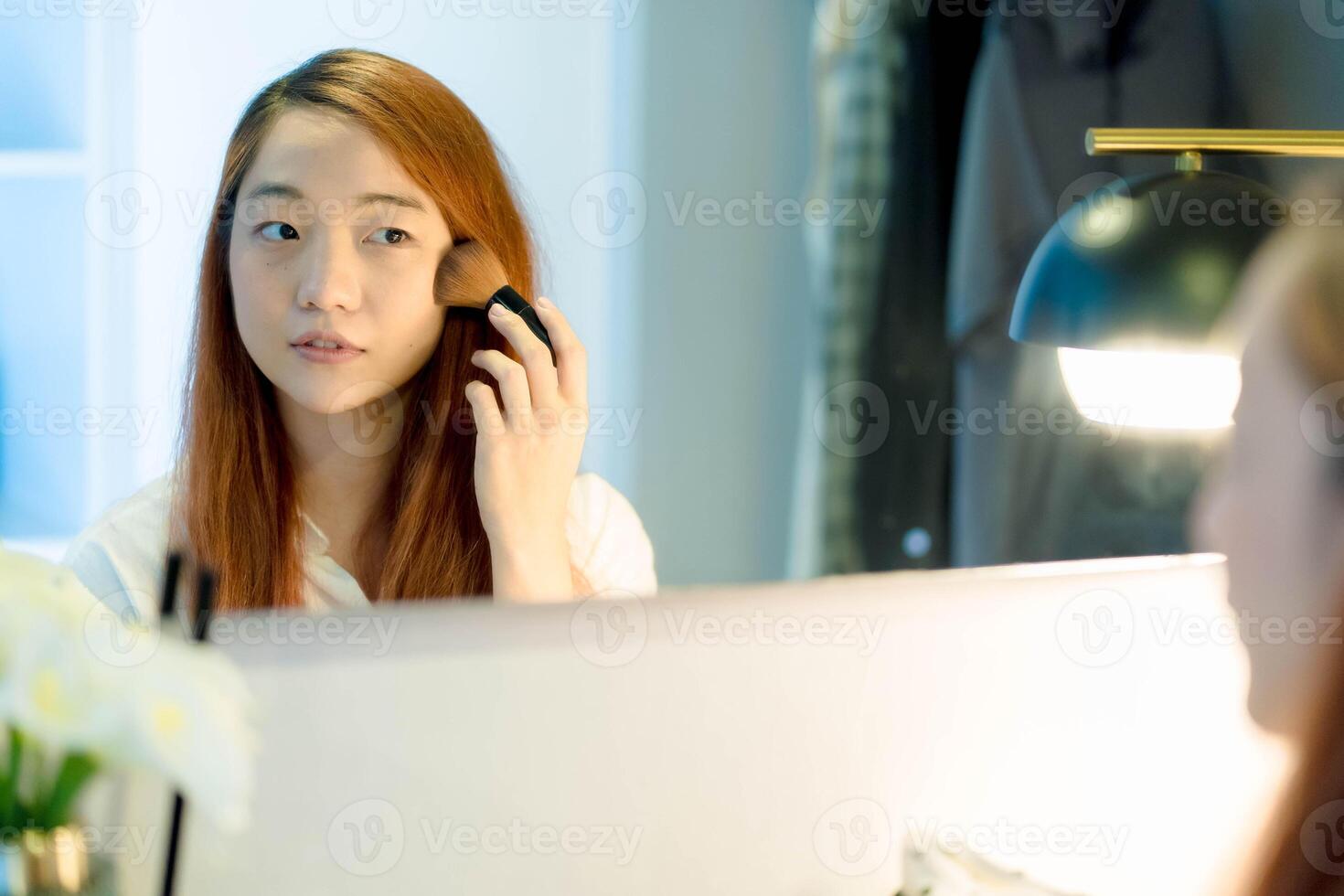 Young Asian woman applying blush with a makeup brush in front of a mirror at home, focusing on her beauty routine in a cozy, well-lit dressing area. photo