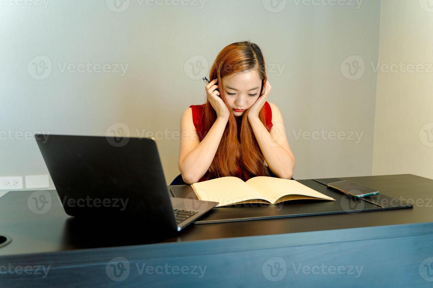Frustrated Asian woman overwhelmed by work, sitting at desk with open notebook, laptop and phone, experiencing stress or burnout while studying or working. photo