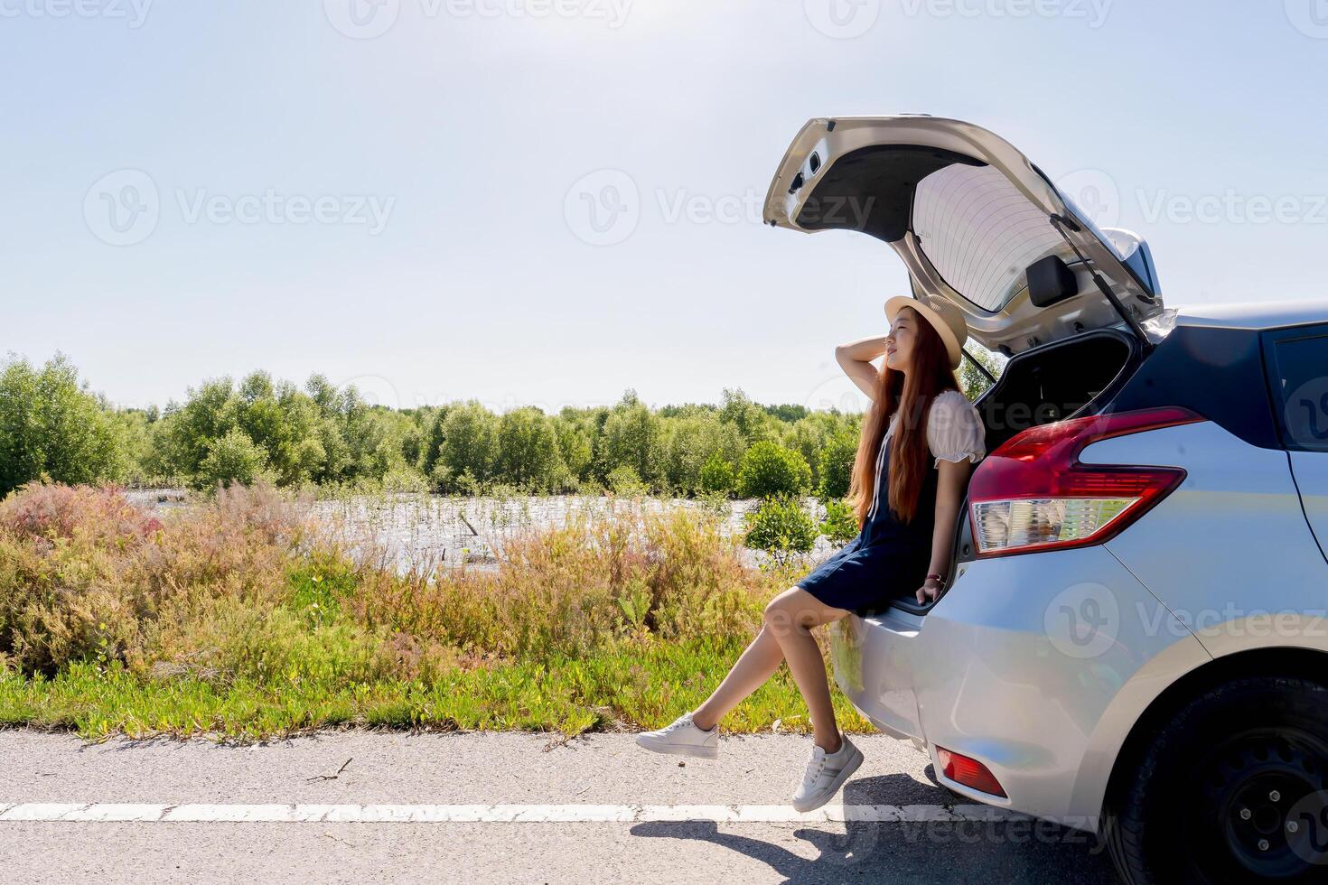 Young female traveler sitting on the back of a hatchback car, enjoying a scenic riverside view on a sunny day during a relaxing road trip adventure. photo