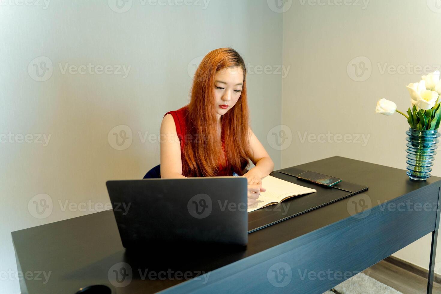 Focused young Asian woman writing notes at a modern black desk with a laptop and flowers in a cozy home office setting, working or studying with intention. photo