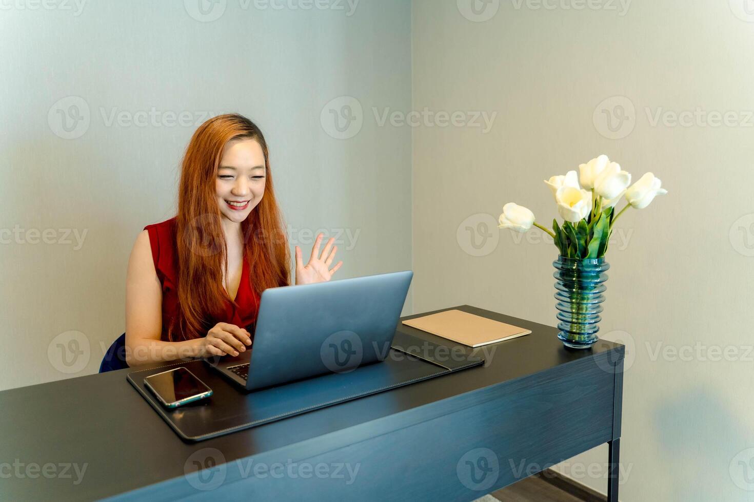 Happy Asian woman waving during call while working remotely from home office with laptop and flower vase on a desk in a modern minimalistic setting. photo