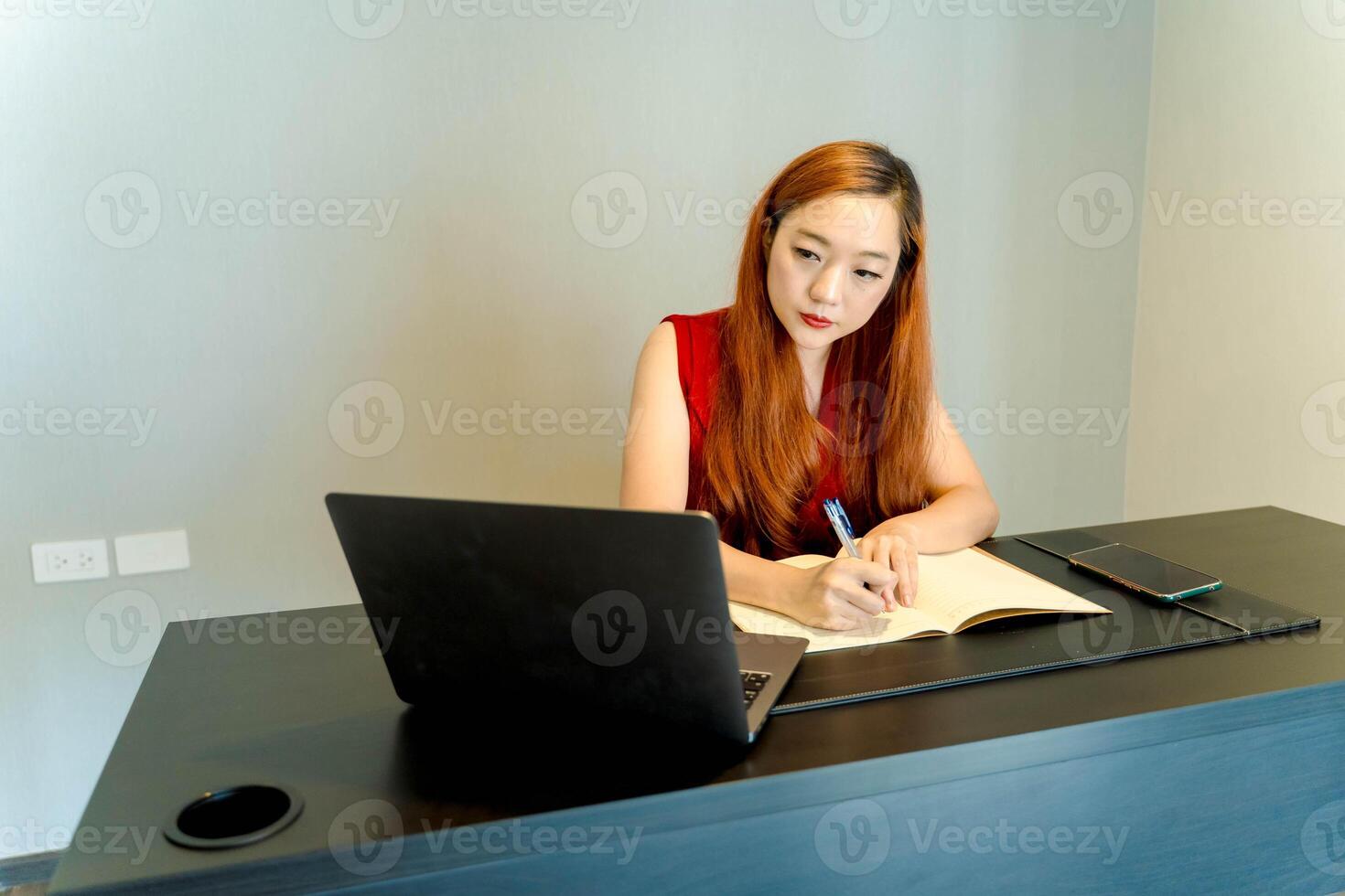 Asian woman writing in a notebook while working from home, focused and engaged at a black desk with laptop and smartphone, minimalist modern workspace. photo