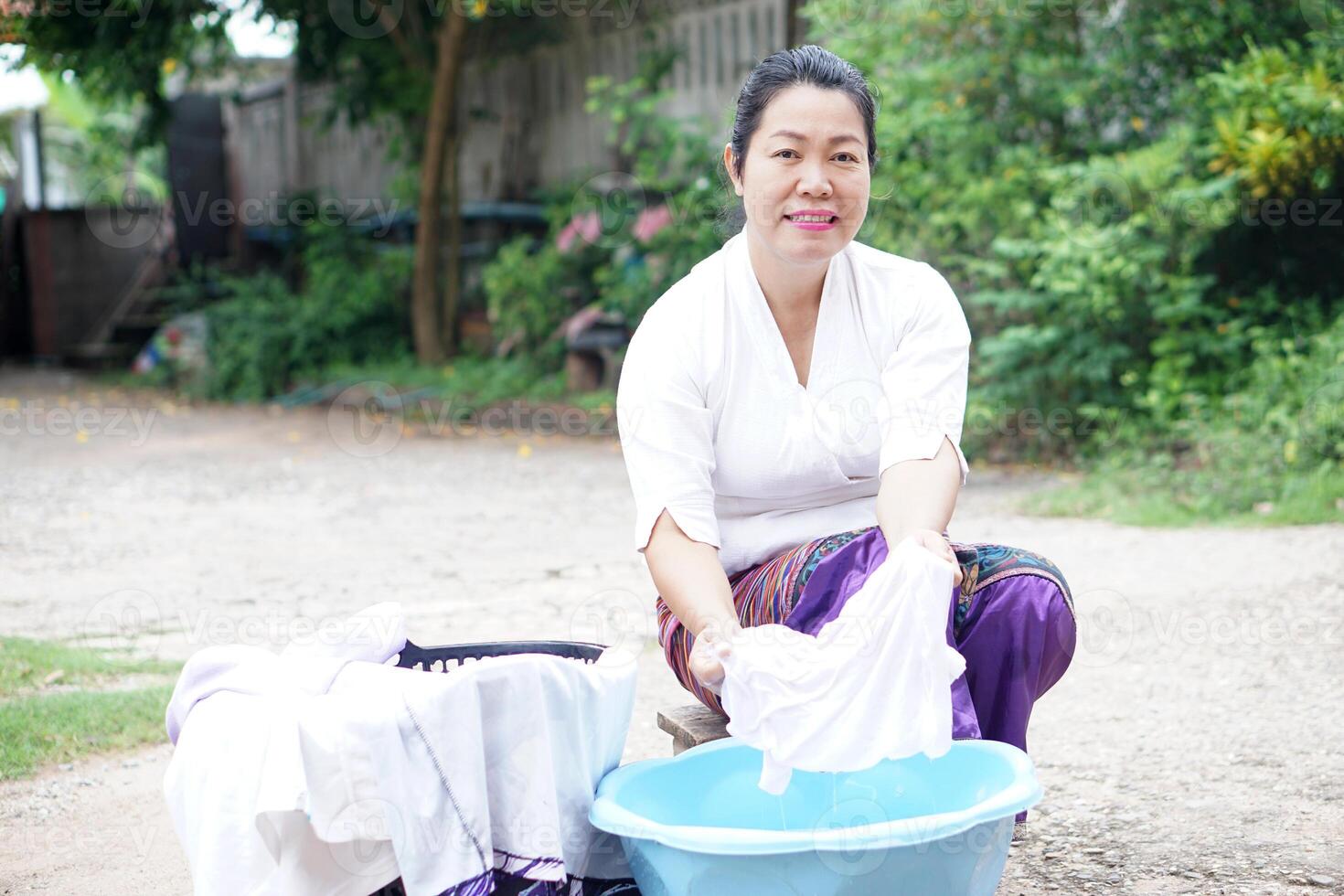 Middle-aged Asian woman in traditional Thai costume hand-washing clothes in a blue basin outdoors, smiling warmly. Rural lifestyle, authentic culture, daily life in Thailand photo