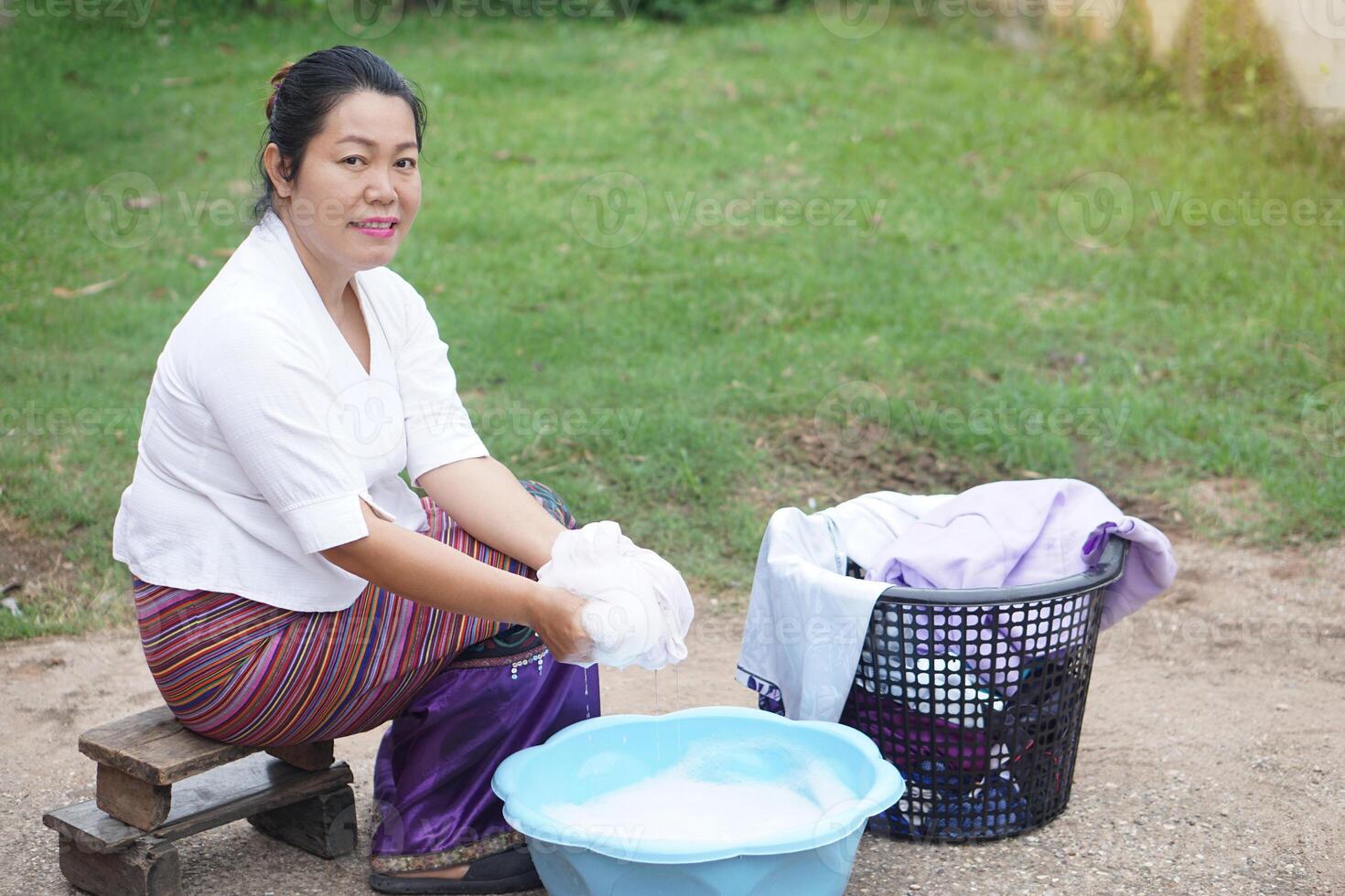 Asian woman in traditional Thai clothing hand-washing laundry in a blue plastic basin, sitting on a wooden stool, with soap suds and a full laundry basket. Rural life in Thailand. photo