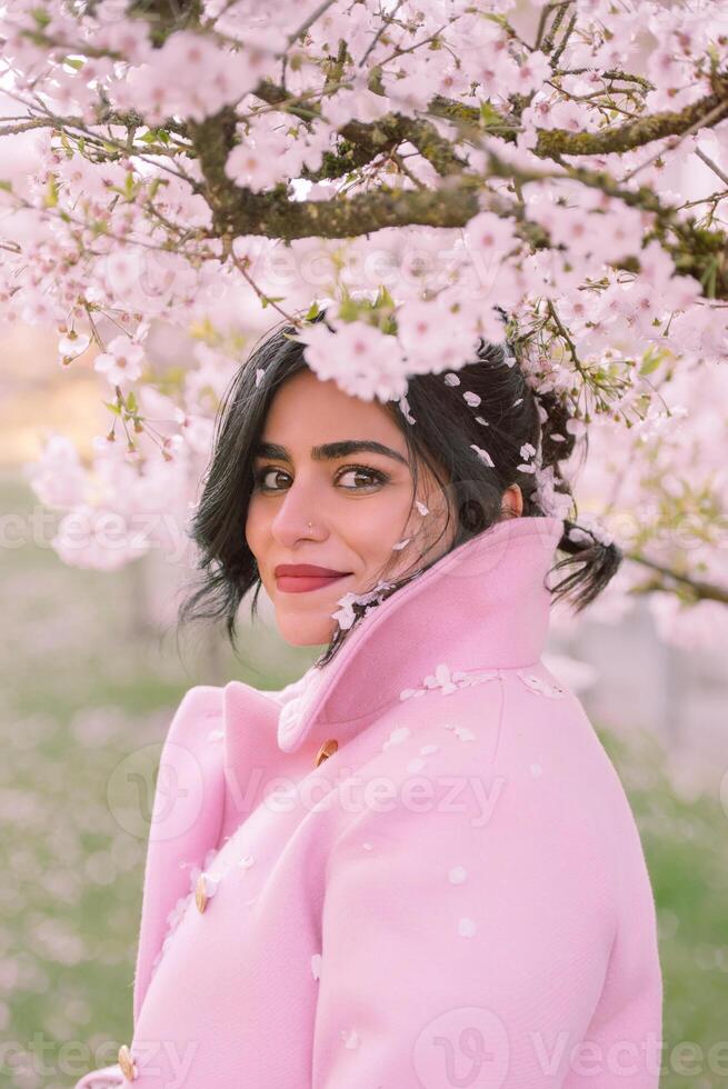 young woman in a soft pink coat standing under blooming cherry blossom trees. photo