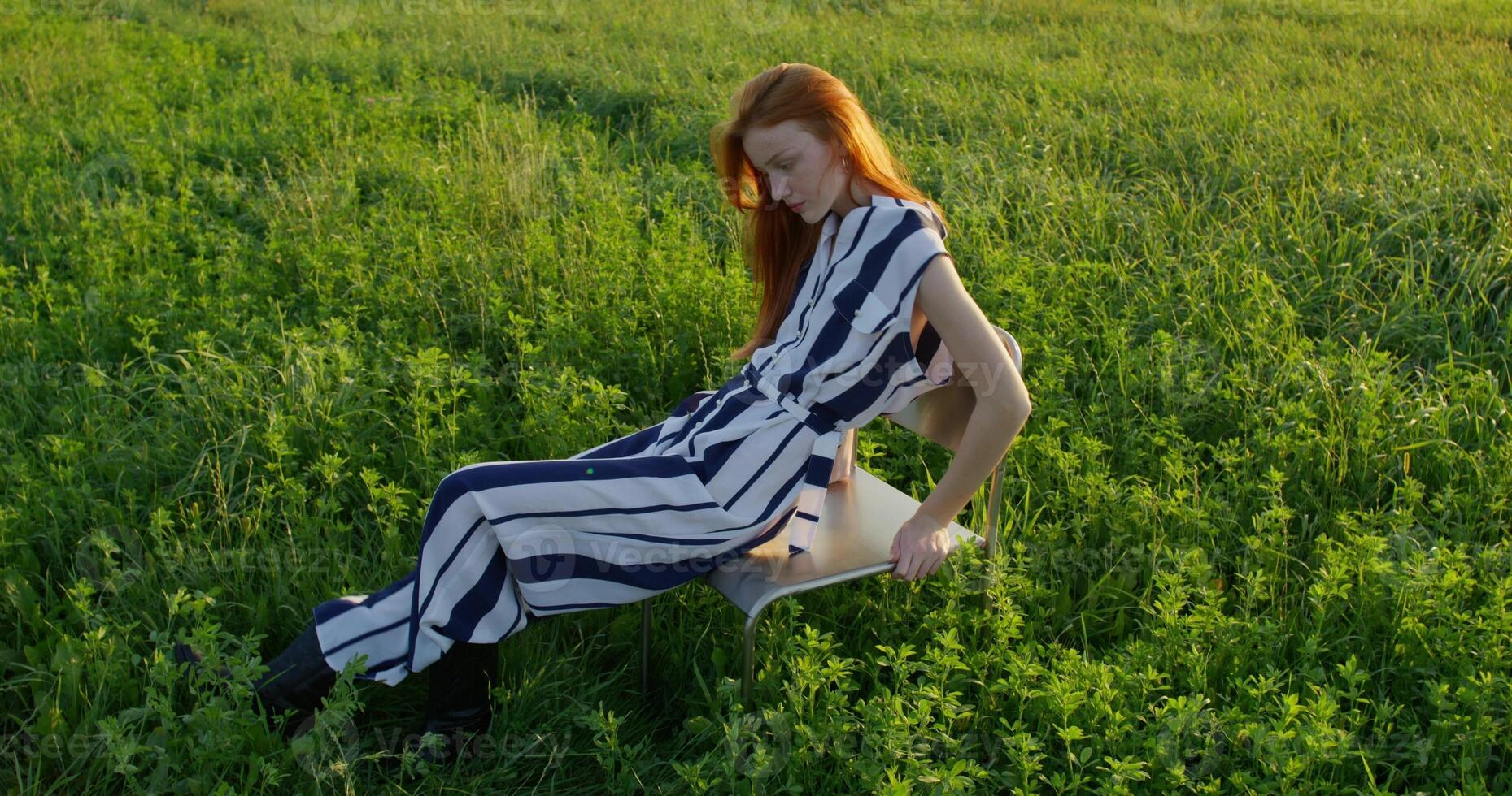 A red-haired woman poses looking at the camera while sitting on a chair in the middle of a field, she smiles. A sunny summer day photo