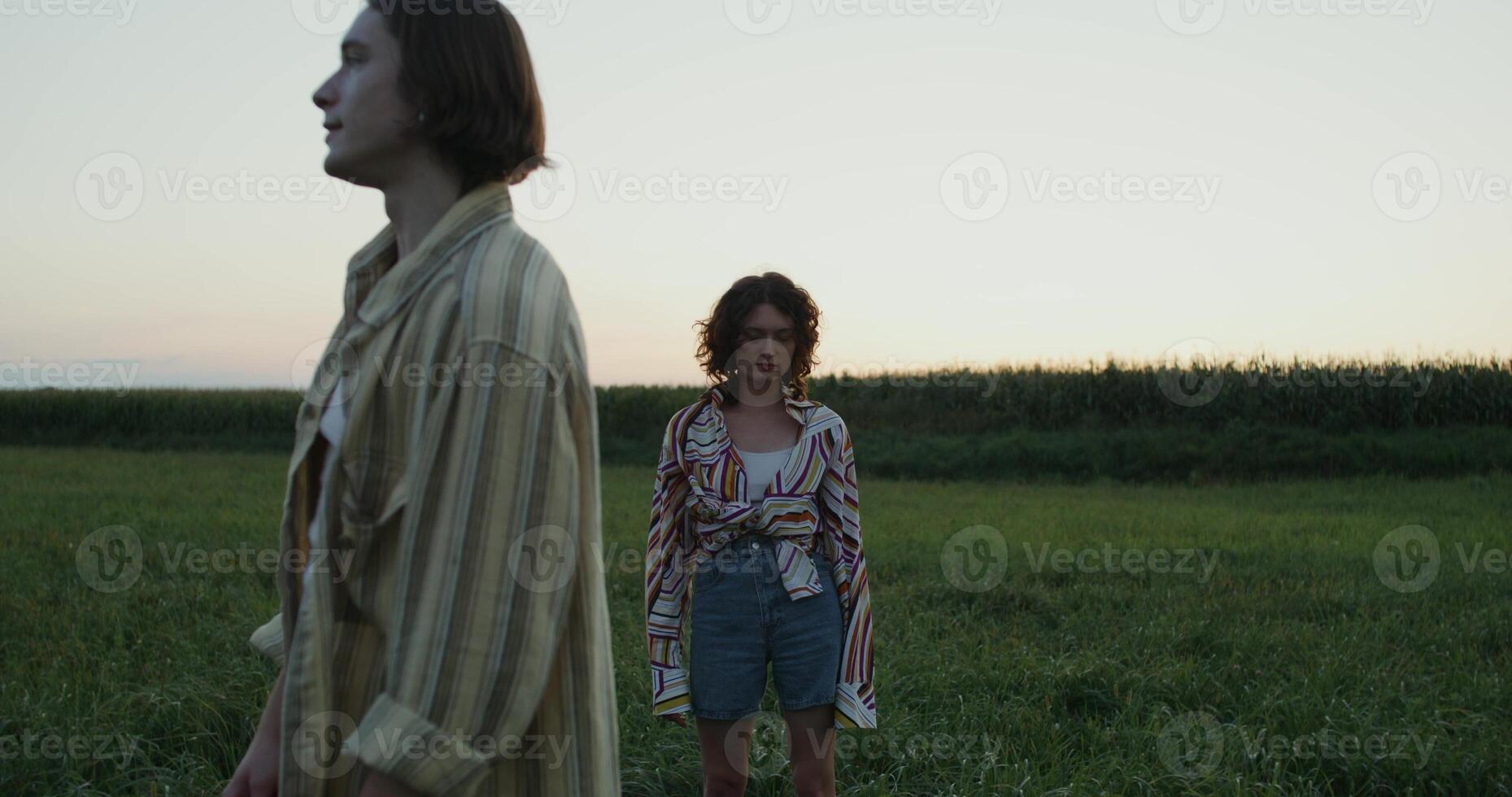 A young man and a woman are standing in a field on a summer evening. The man looks into the distance. A woman looks at the camera photo