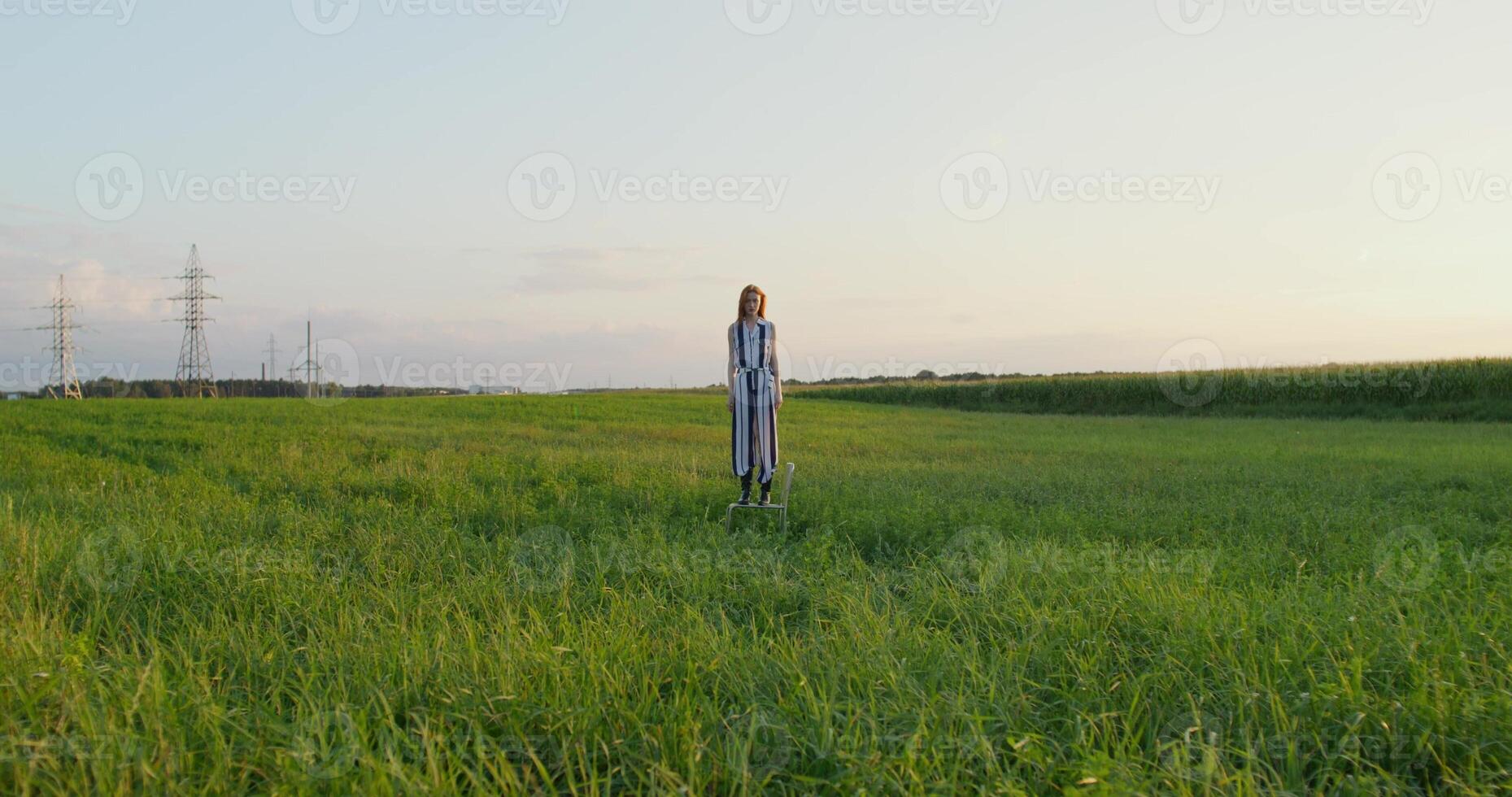 A red-haired woman poses standing on a chair in the middle of a field at sunset with a serious look photo