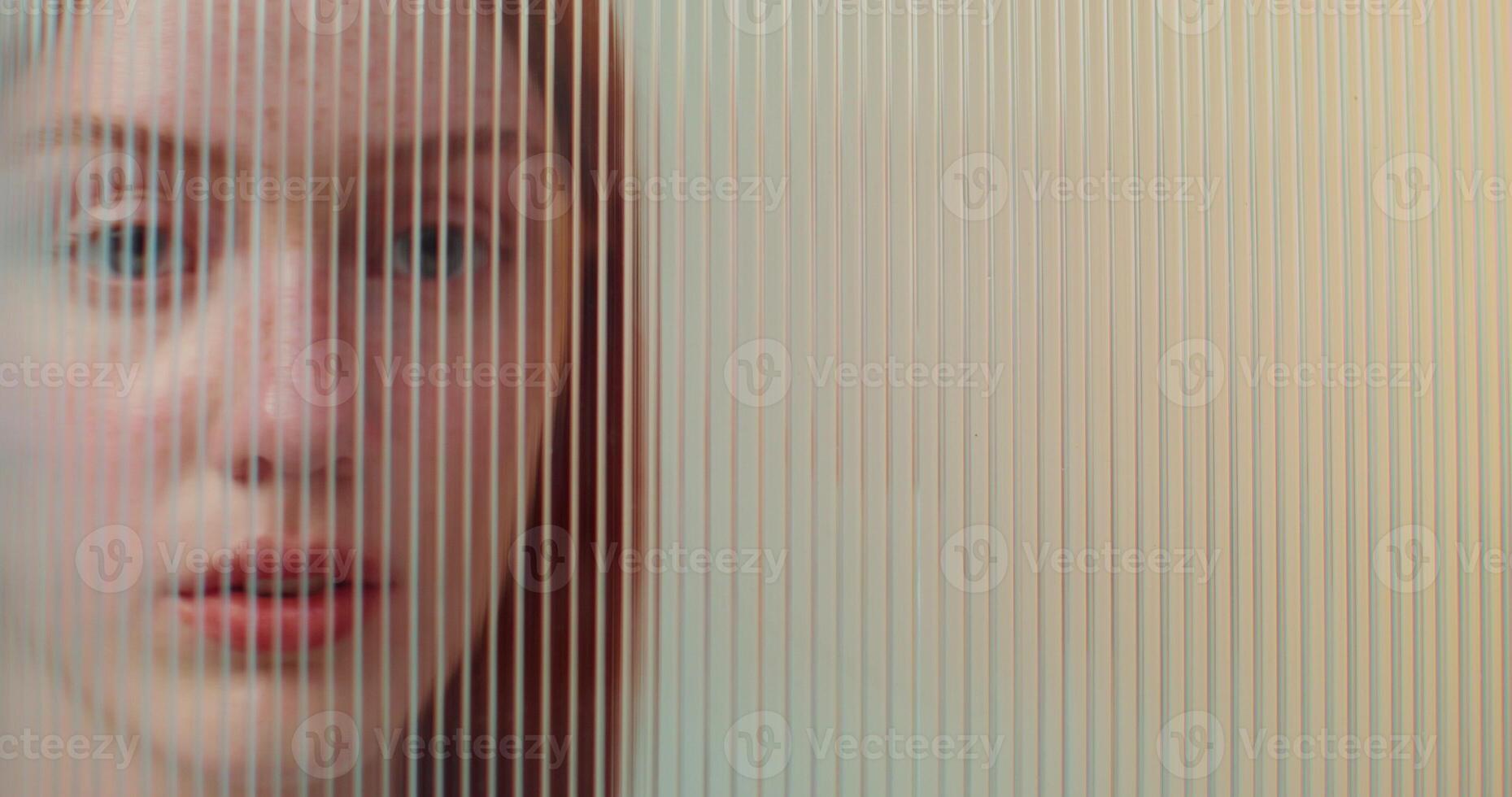A red-haired woman with freckles looks at the camera with a serious look standing behind striped plexiglass in the studio photo