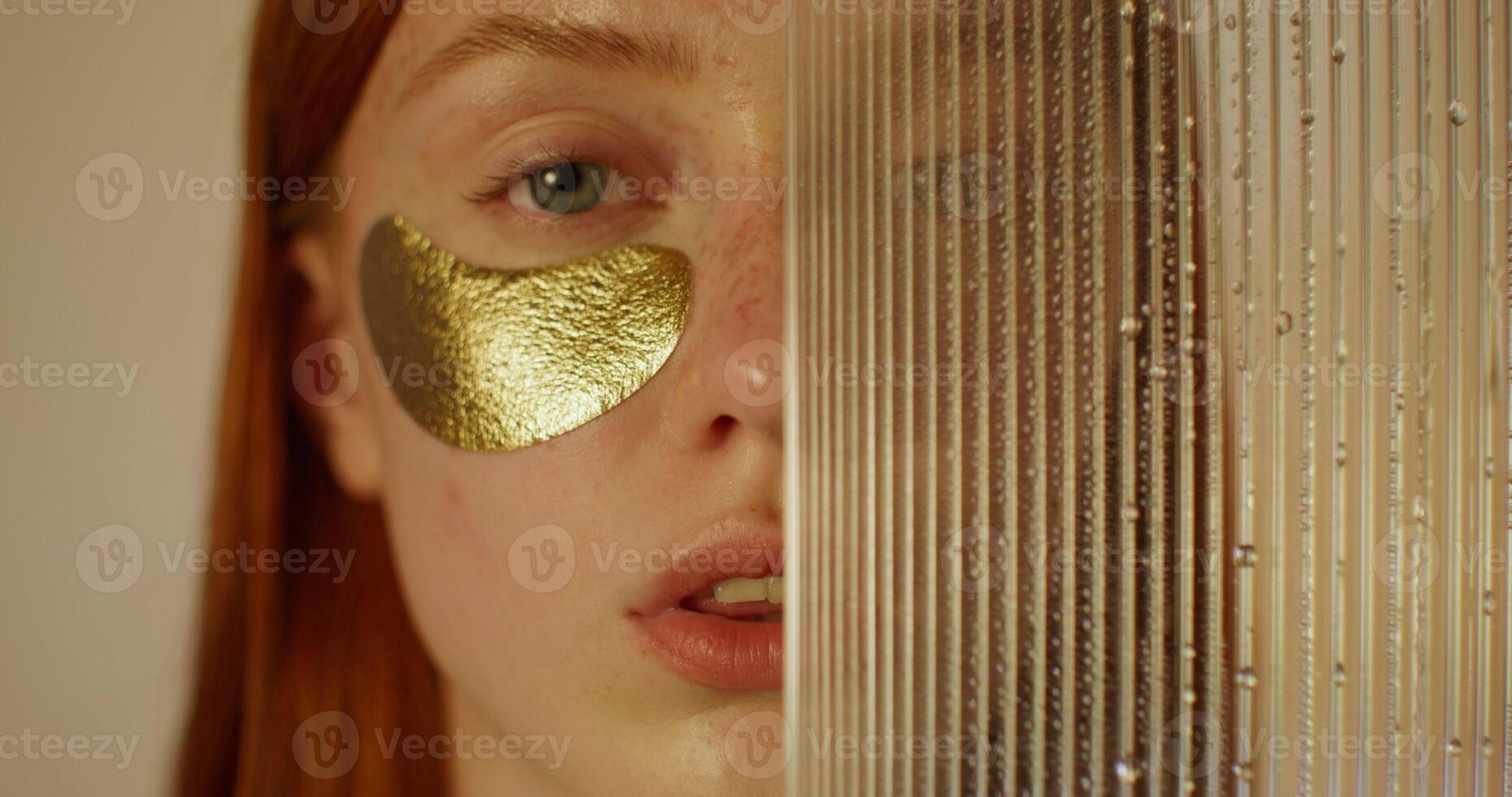 A woman with an eye patch looks at the camera with a serious look. Half of her face is covered with glass with water running down it. Studio photo