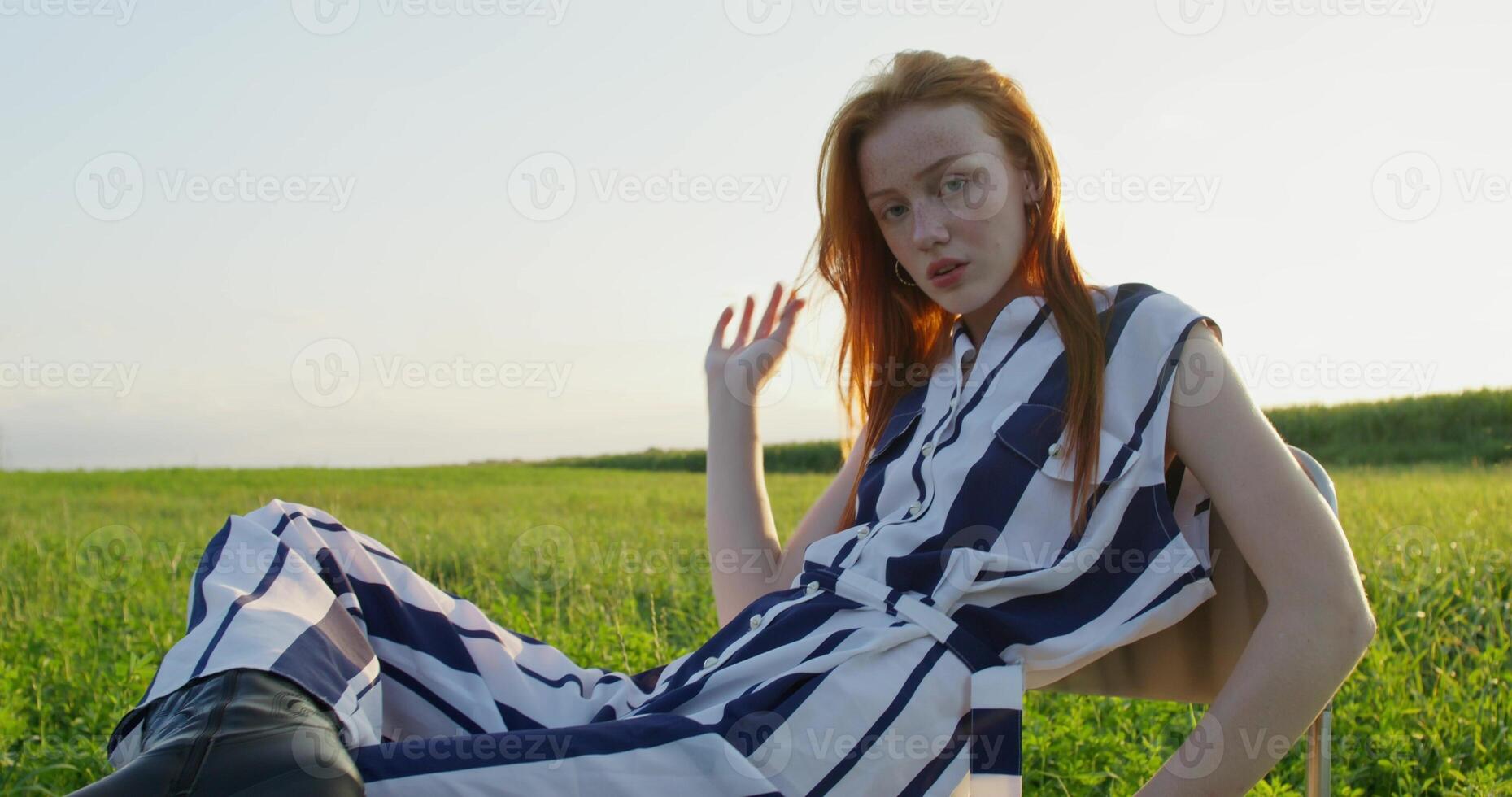 A red-haired woman poses looking at the camera with a calm look while sitting on a chair in the middle of a field. A sunny summer day photo