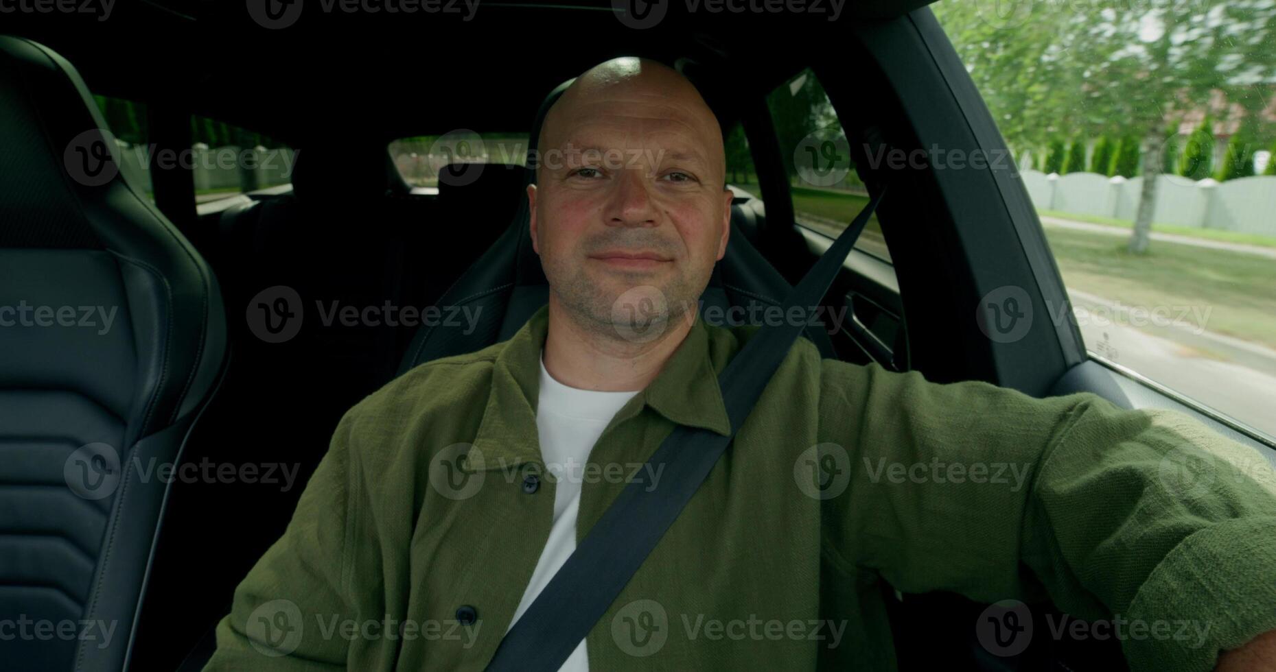 The man is driving the car, he is wearing a seat belt. He's smiling. Front view inside the car. Summer day photo