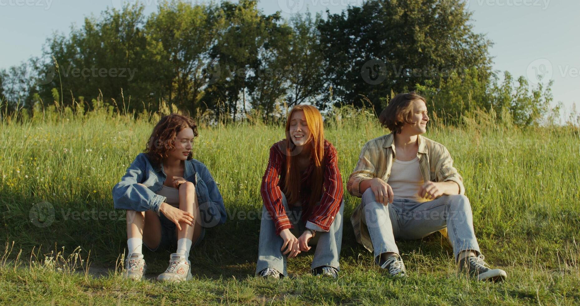 Three young people are talking sitting on the ground in a field. They look tired and happy. A sunny summer day photo