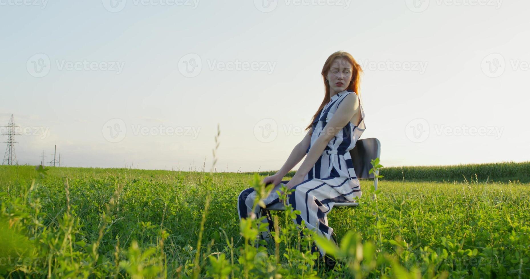 A red-haired woman poses sitting on a chair in the middle of a field. A sunny summer day photo