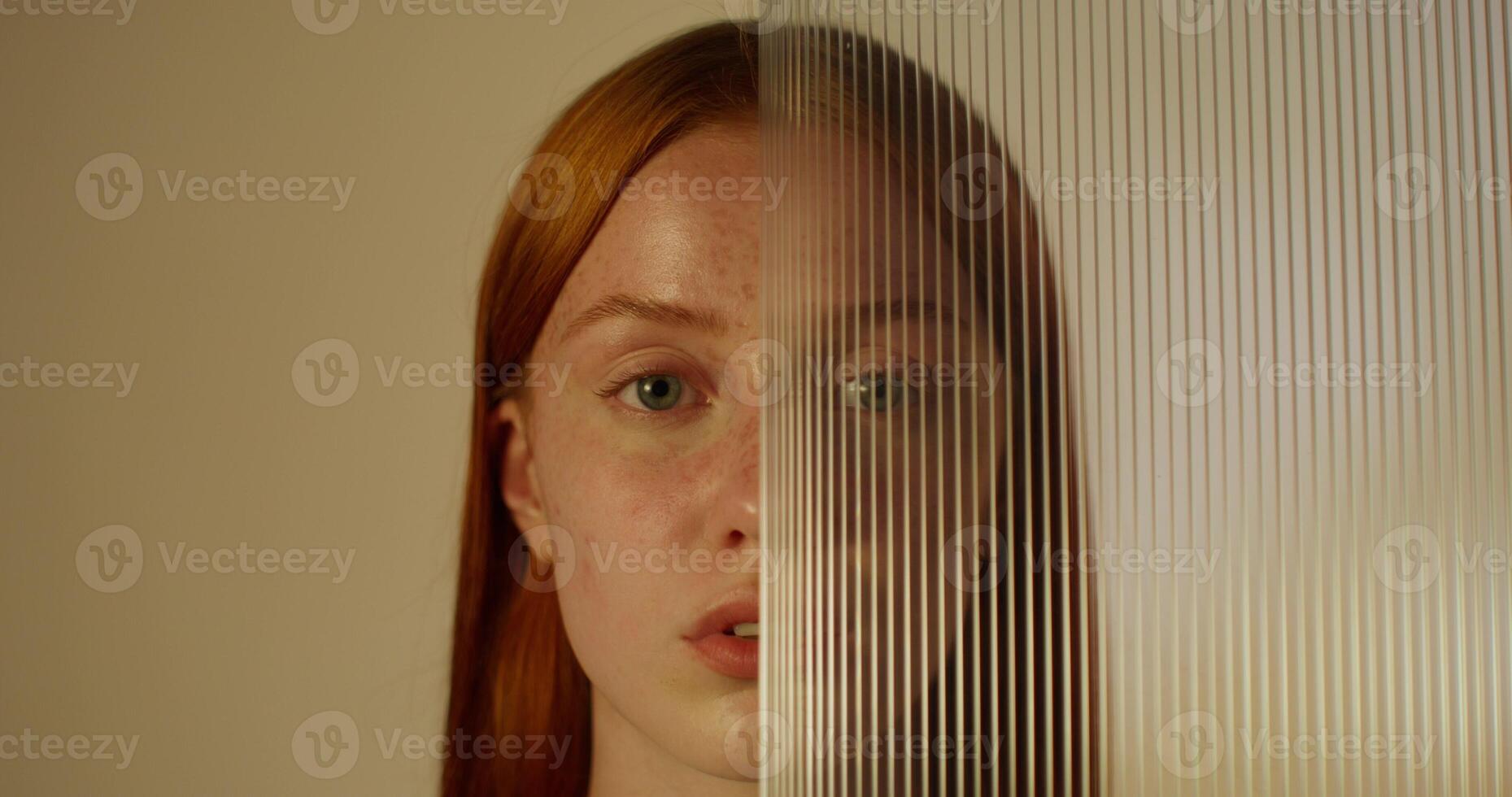 A red-haired woman with freckles looks at the camera with a serious look standing on a studio background. Half of her face is covered with plexiglass photo