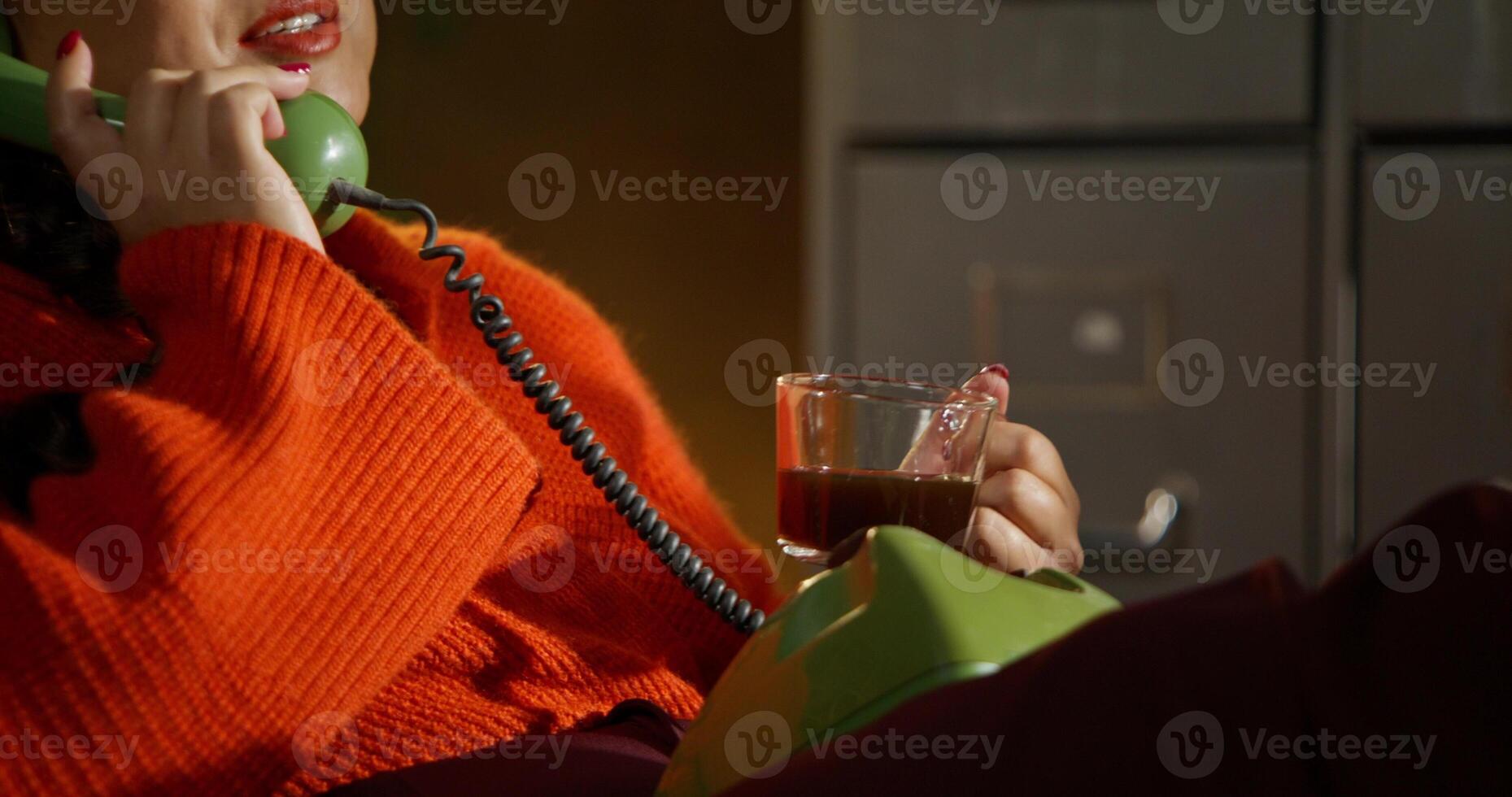 A woman is talking on a wired phone and drinking coffee while sitting in a retro office photo